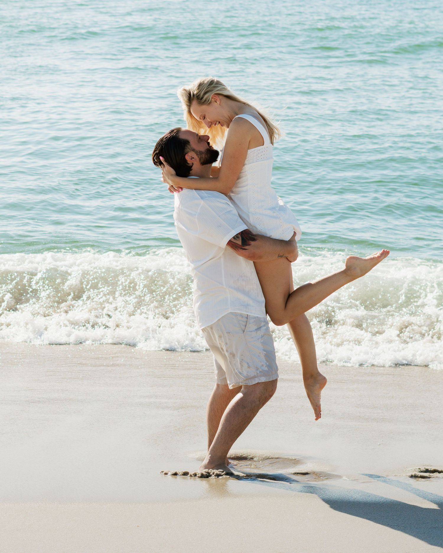 Couple in white outfits embracing and kissing on a sunny beach with ocean waves in the background.