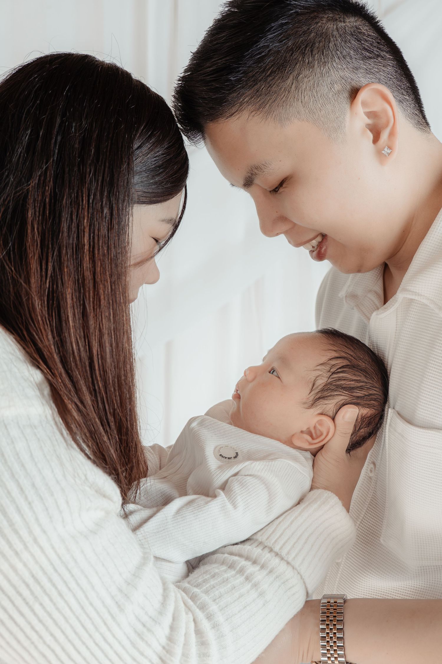 Young Asian parents smiling lovingly at their newborn baby, dressed in white, sharing a tender family moment.