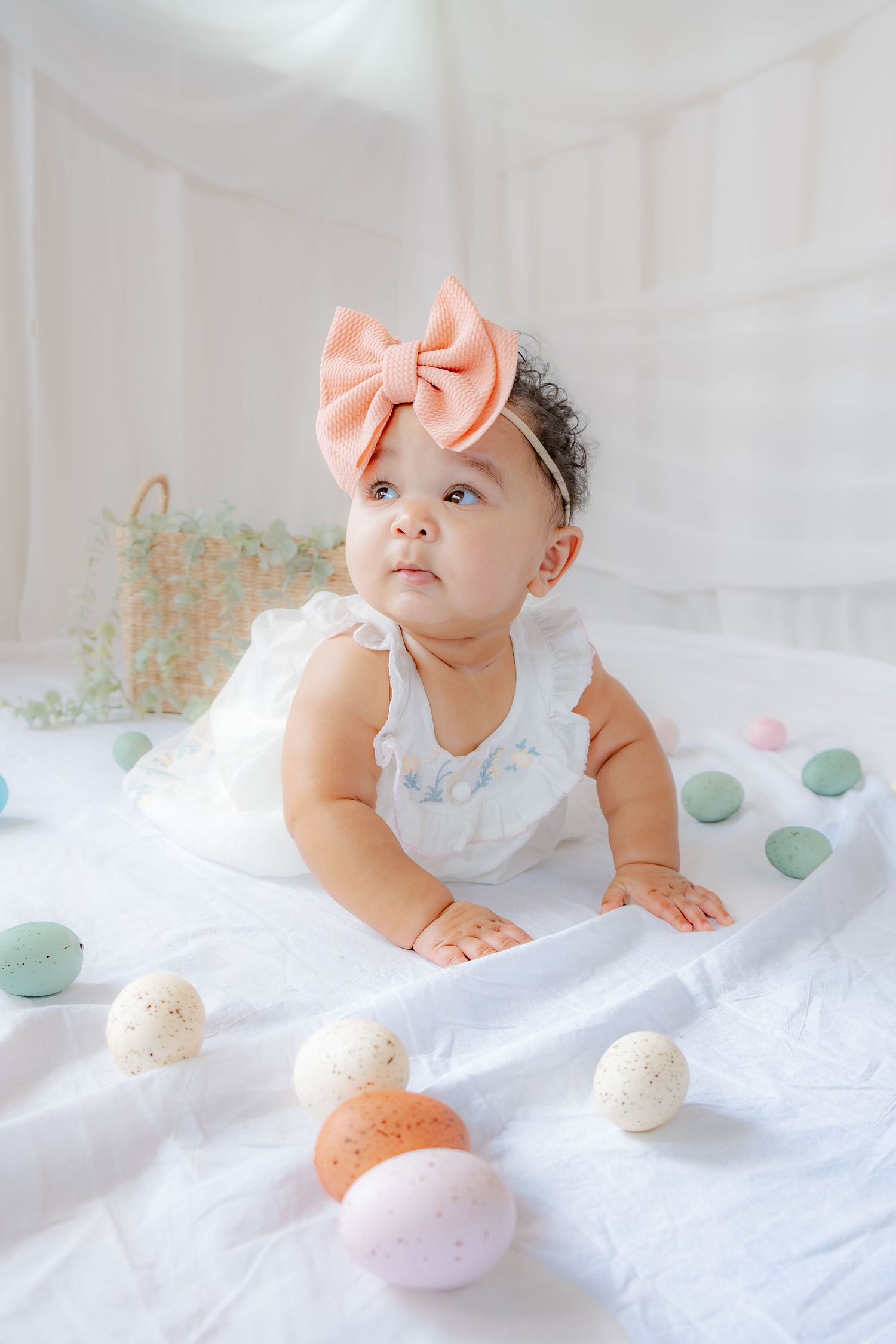 An adorable baby girl in a white dress and peach bow headband lies surrounded by colorful pastel Easter eggs.