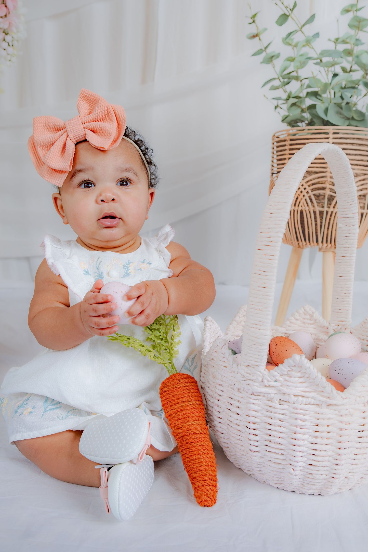 Baby girl with pink bow headband sitting beside wicker Easter basket with carrots.