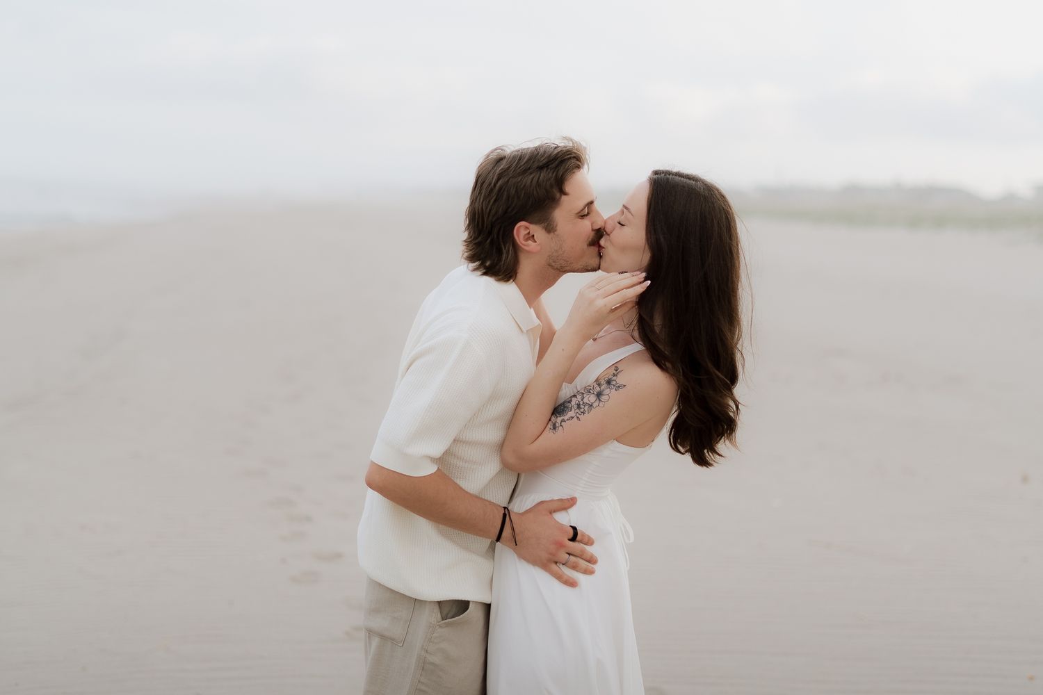 Couple sharing a romantic kiss on a sandy beach, woman in white dress with floral tattoo, misty coastal backdrop.