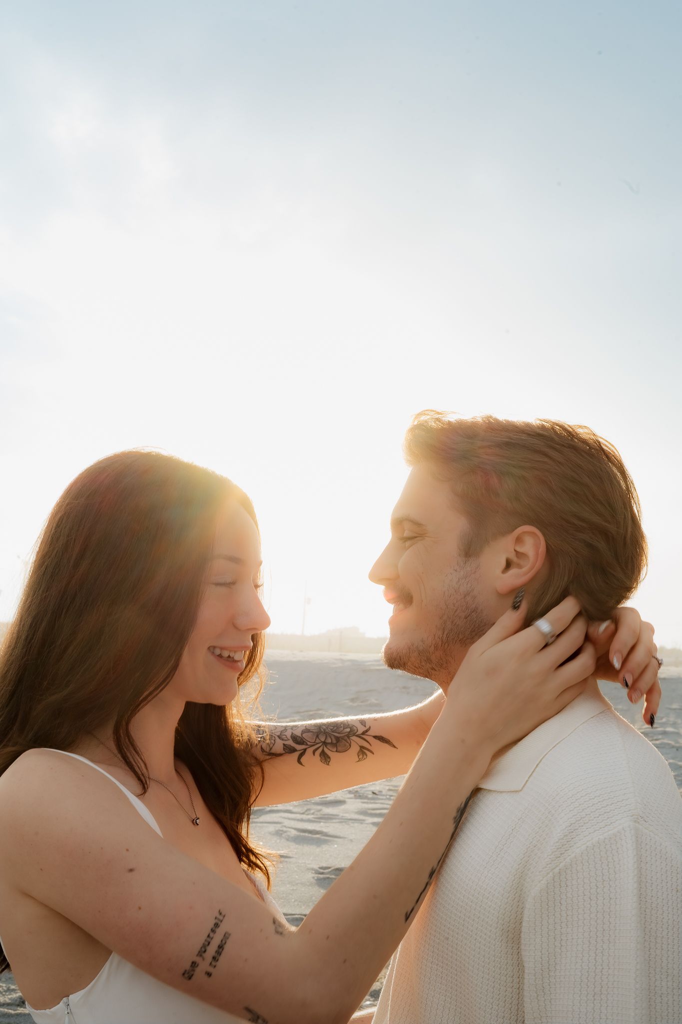 Couple smiling at each other on a sunny beach, woman with tattoos embracing man in warm golden light.