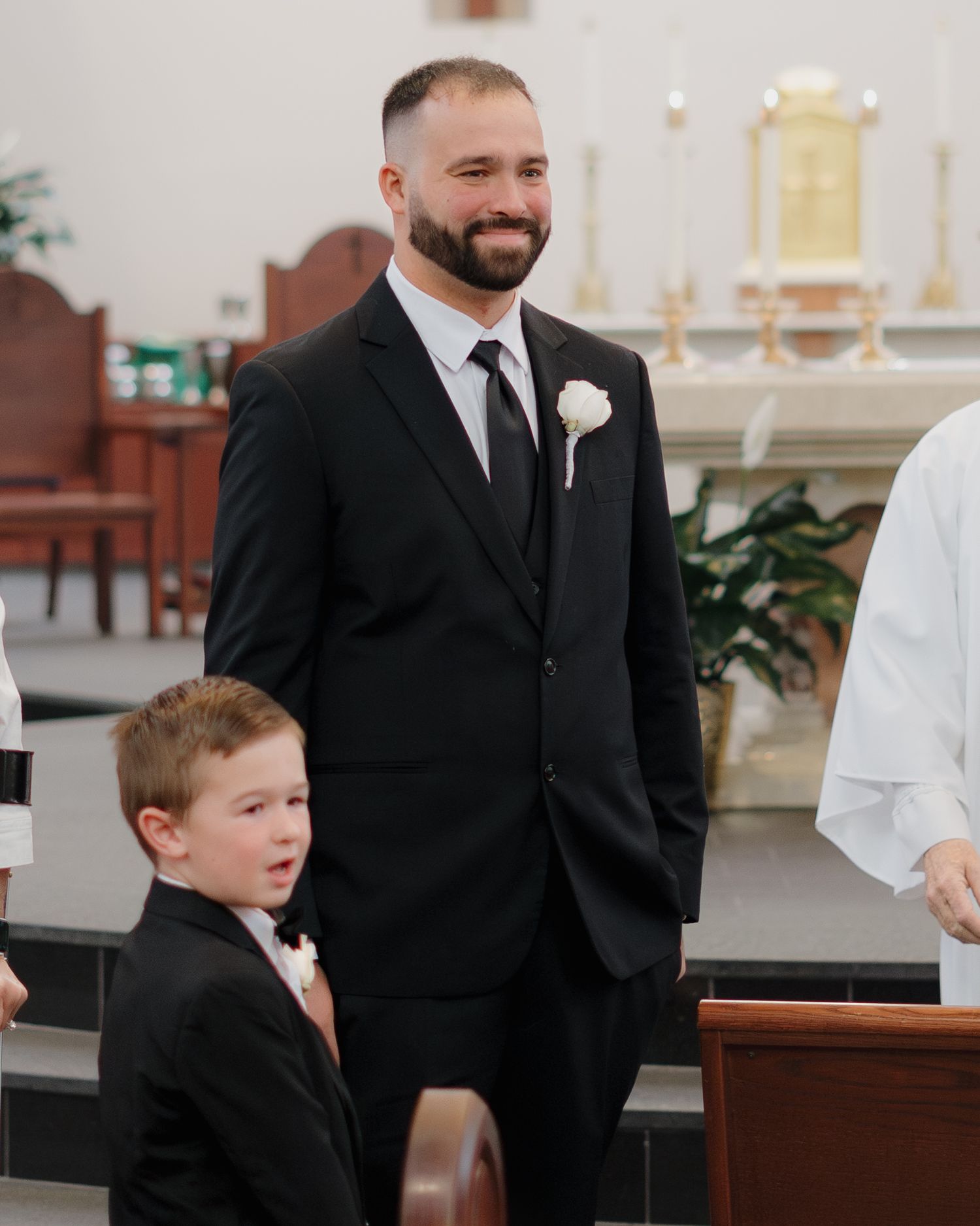 A smiling groom in a black suit with white boutonniere stands beside a young boy in a church wedding ceremony.