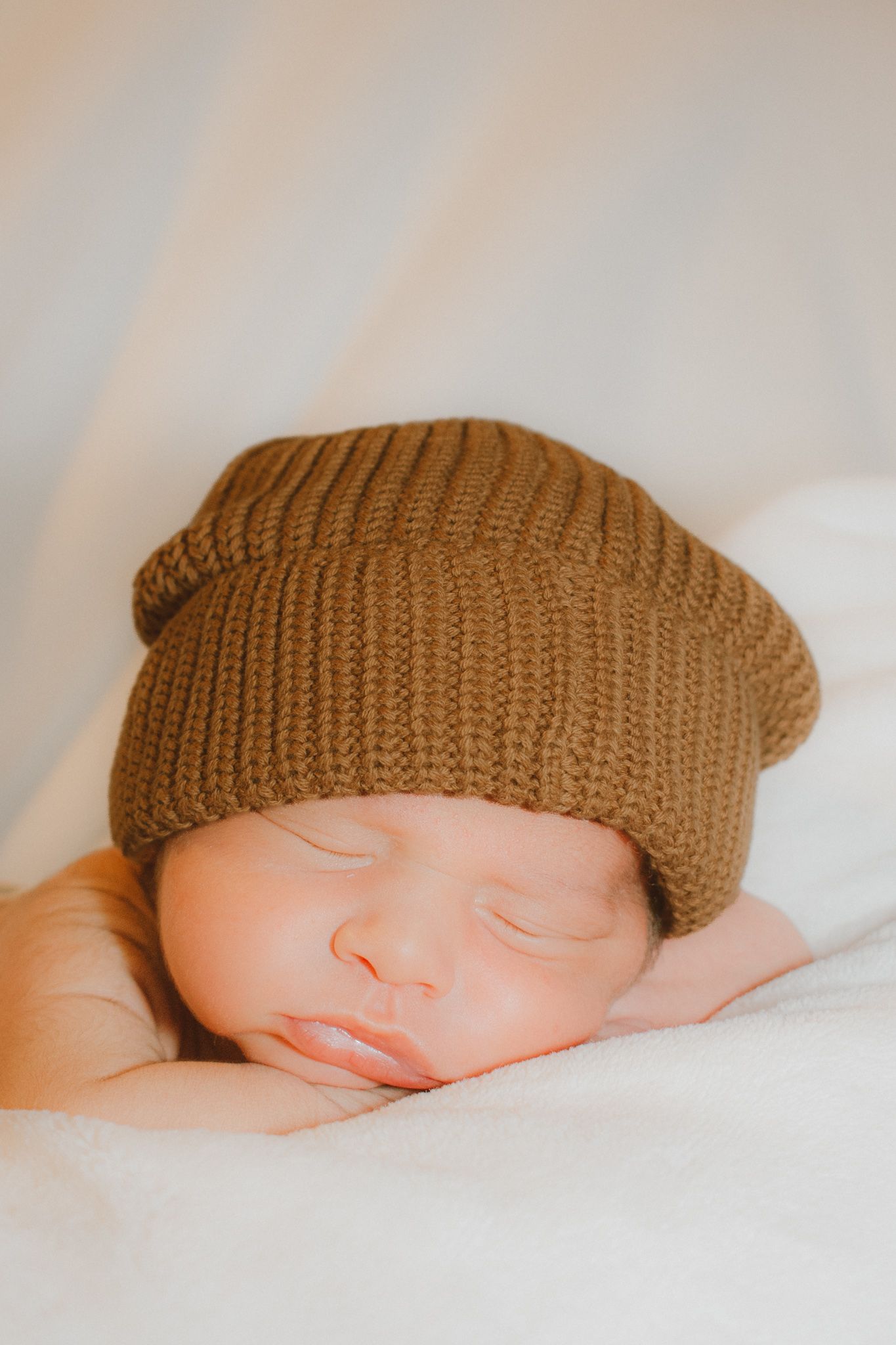 Sleeping newborn baby wearing an oversized brown knit beanie hat on white background.