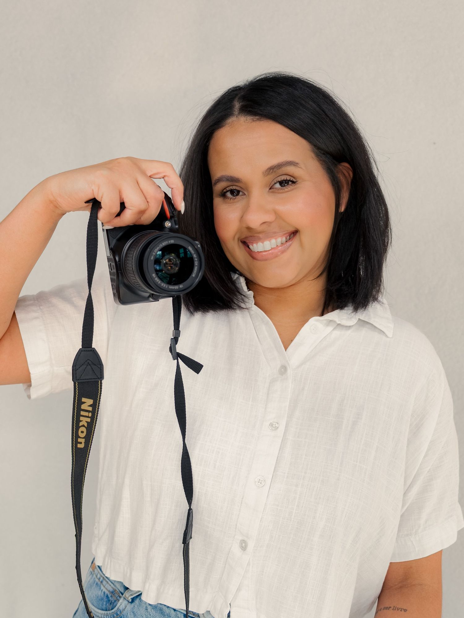 Smiling woman in white linen shirt holds Nikon DSLR camera against neutral gray background for photographer portrait.