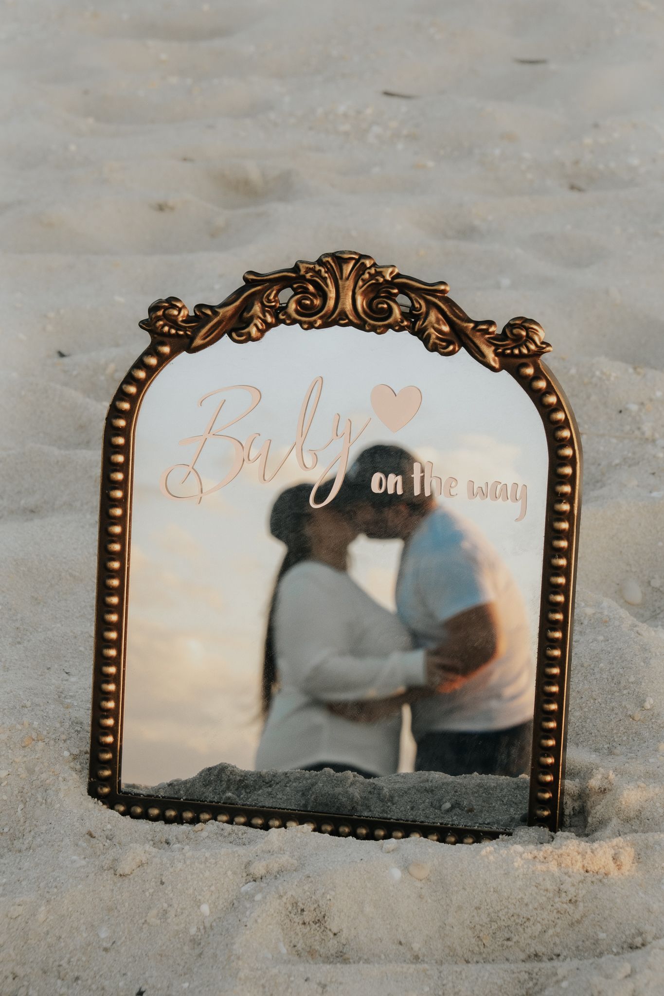 Ornate gold mirror in sand displaying Baby on the Way text reflecting kissing pregnant couple at beach.