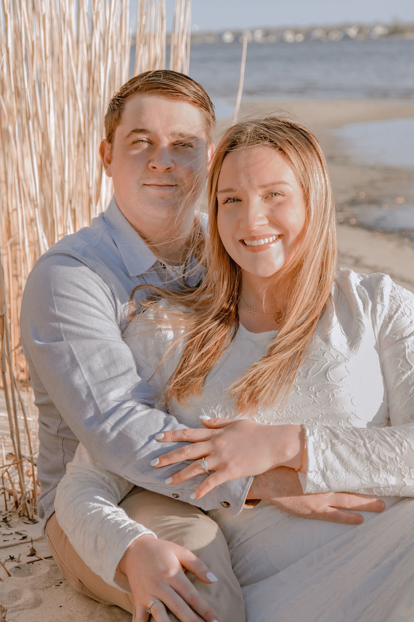 Couple sitting on sandy beach embracing, woman in white lace dress and man in blue shirt, smiling at camera.