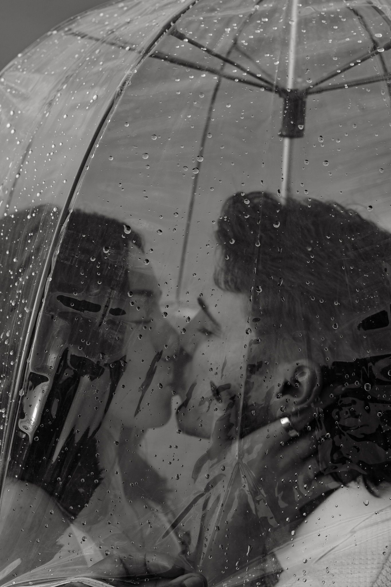 Black and white photo of a couple sharing a romantic moment under a clear umbrella in the rain.