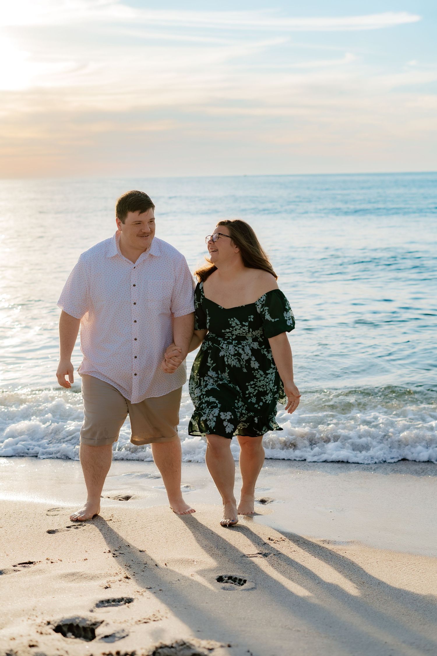 Couple walking hand-in-hand along sandy beach at sunset, waves lapping shore, man in white shirt and woman in floral dress.