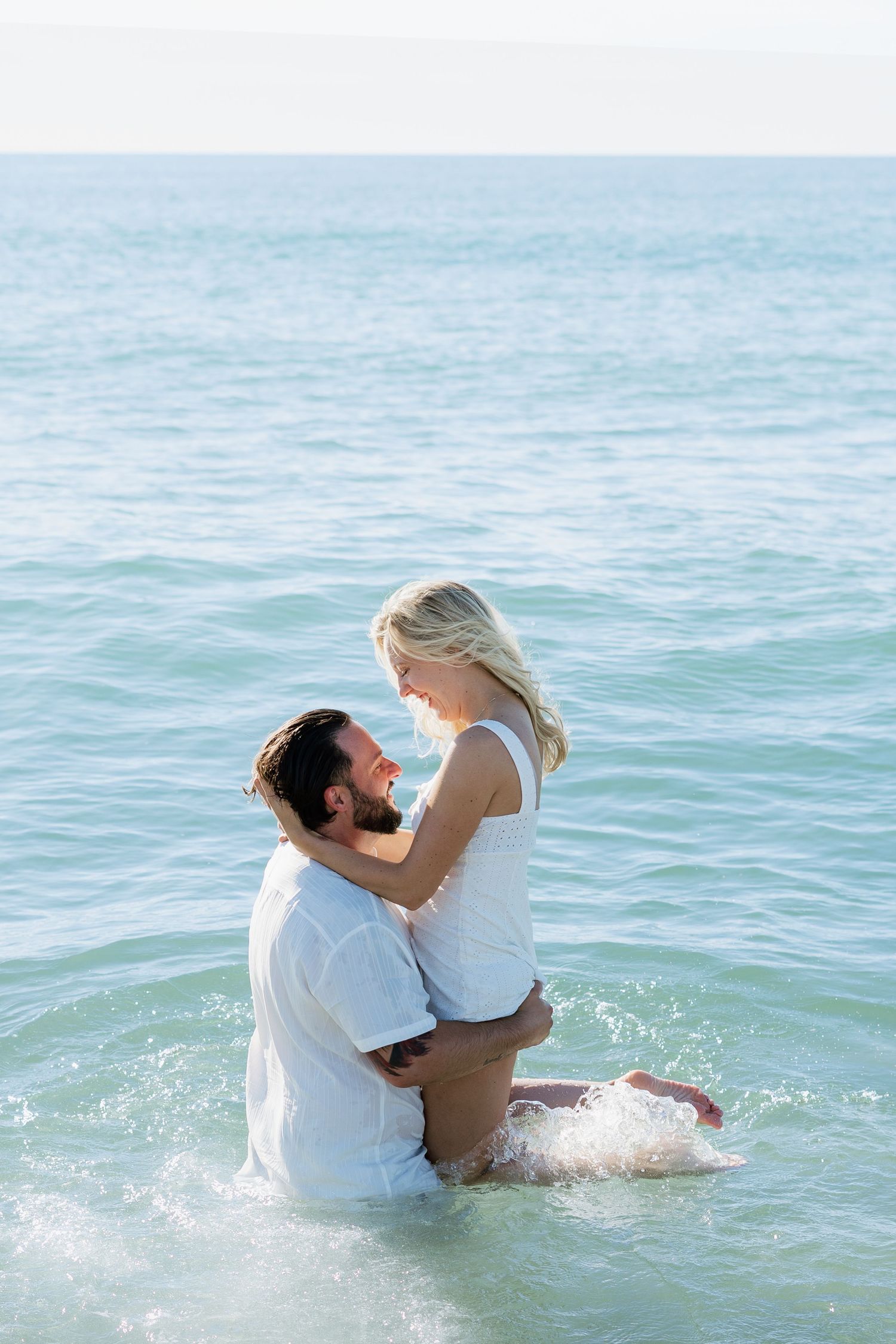 A couple in white clothes embraces romantically while standing in the ocean waves on a sunny day.