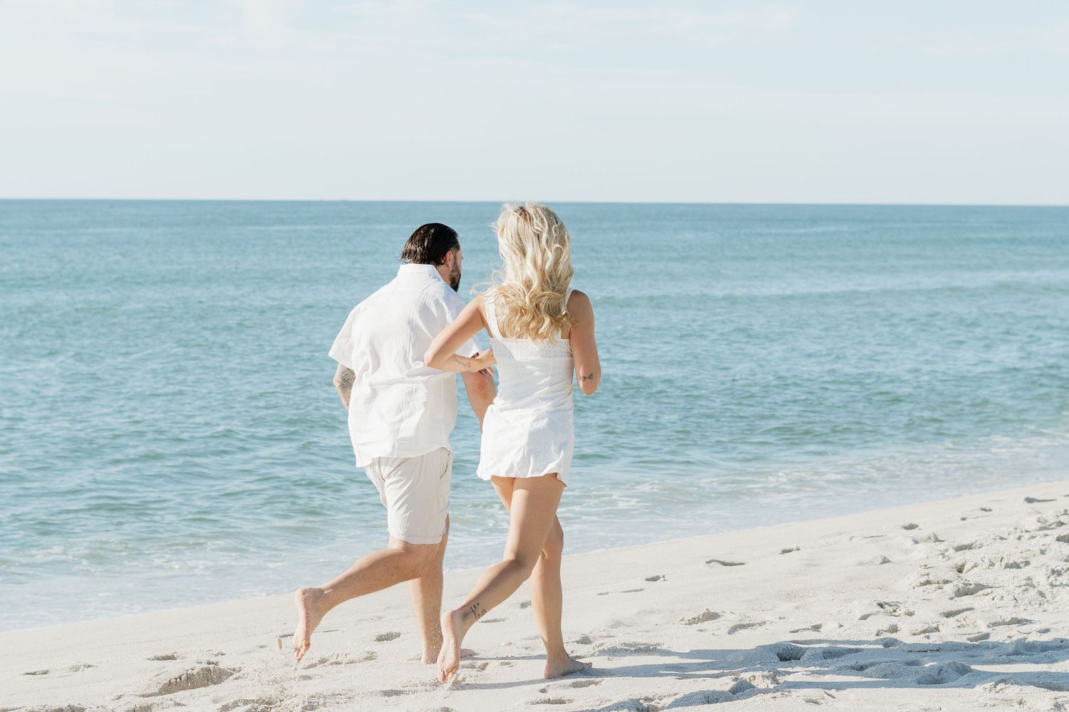 Couple in white outfits running barefoot on a sunny white sand beach with calm blue ocean waves.