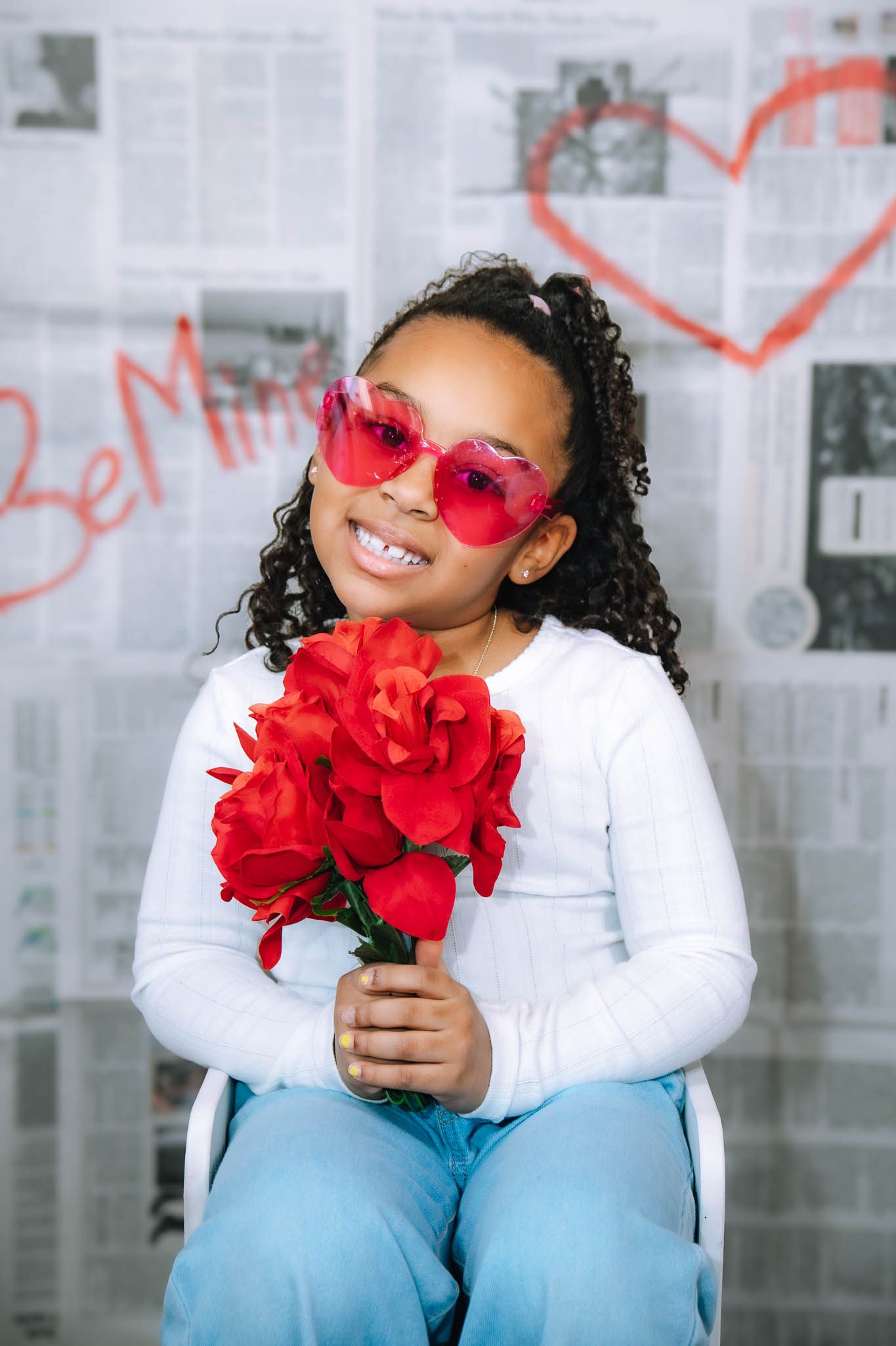 Young girl in pink heart sunglasses holding red flowers in front of newspaper backdrop with Valentine's Day decorations.