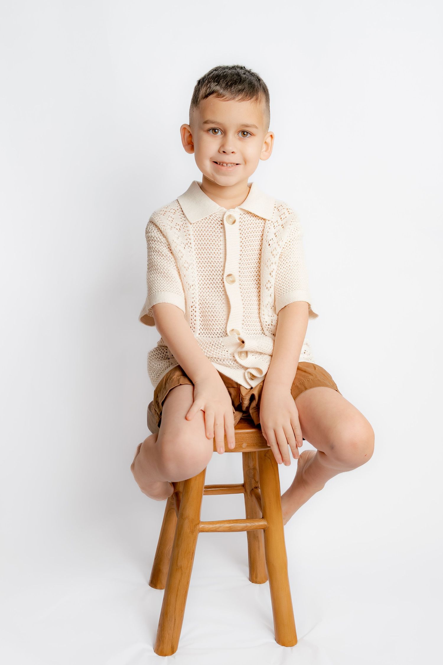 A smiling boy in a cream knit shirt and brown shorts sits confidently on a wooden stool against white backdrop.