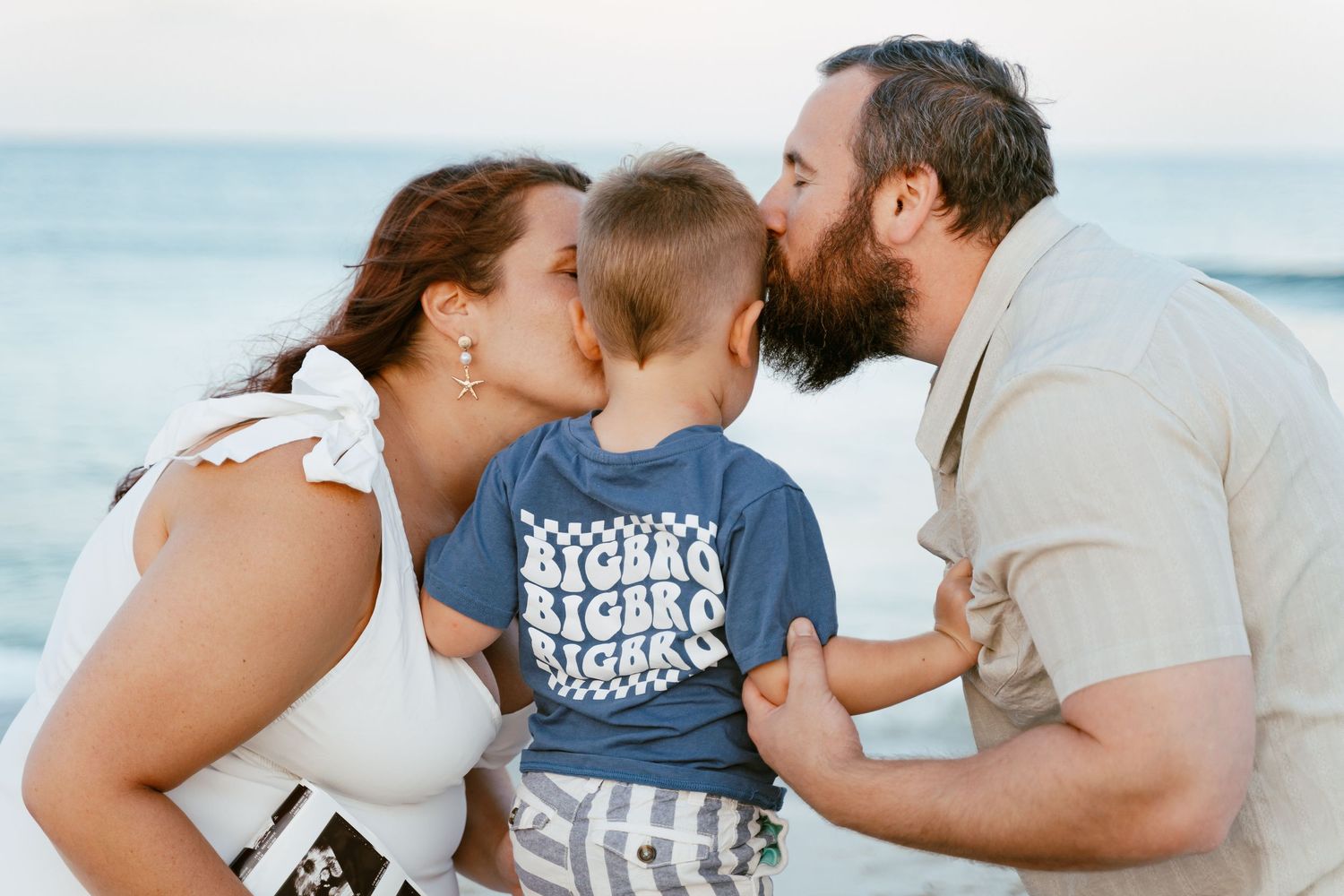 Parents kissing baby boy on cheeks at beach, family portrait by ocean.