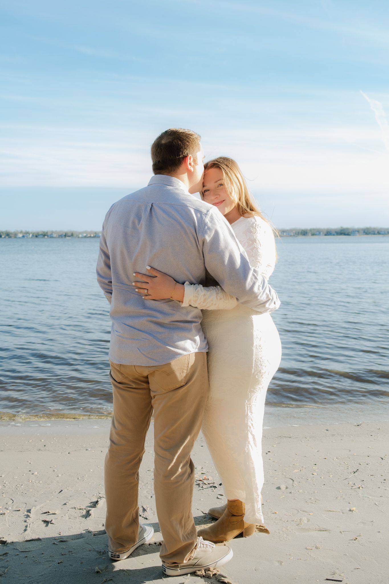 Couple embracing on a sandy beach near calm blue water, woman in white dress smiling, man in light blue shirt and khaki pants.