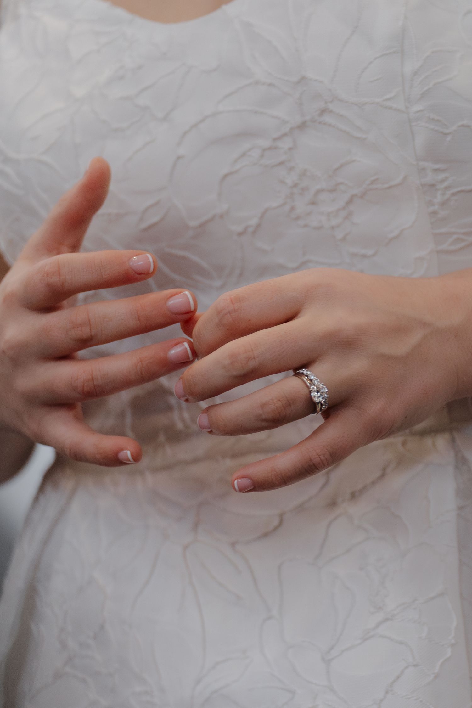 Close-up of bride's hands displaying a sparkling diamond wedding ring set against a white lace bridal gown.