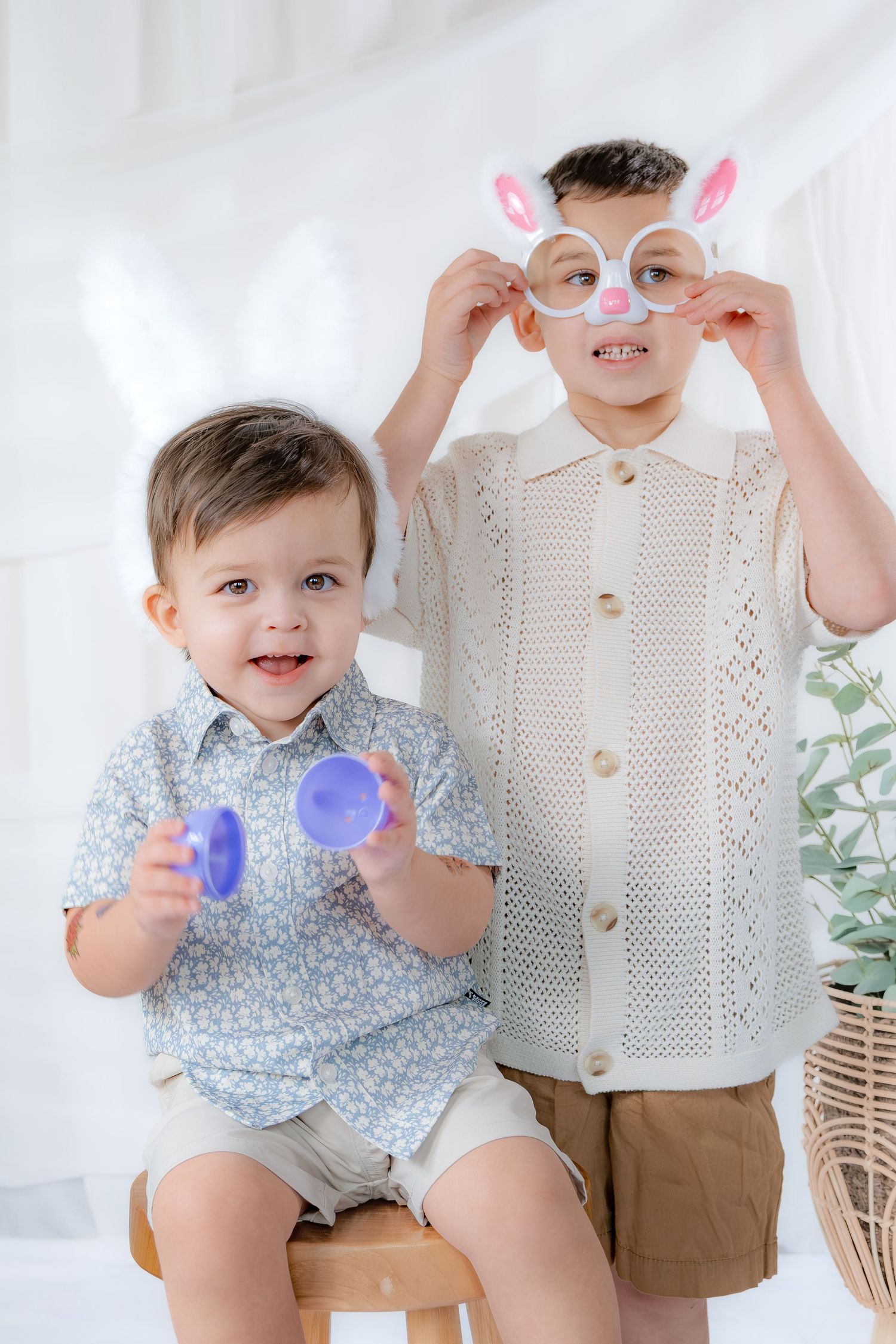 Two young boys in Easter bunny accessories holding colorful Easter eggs indoors.