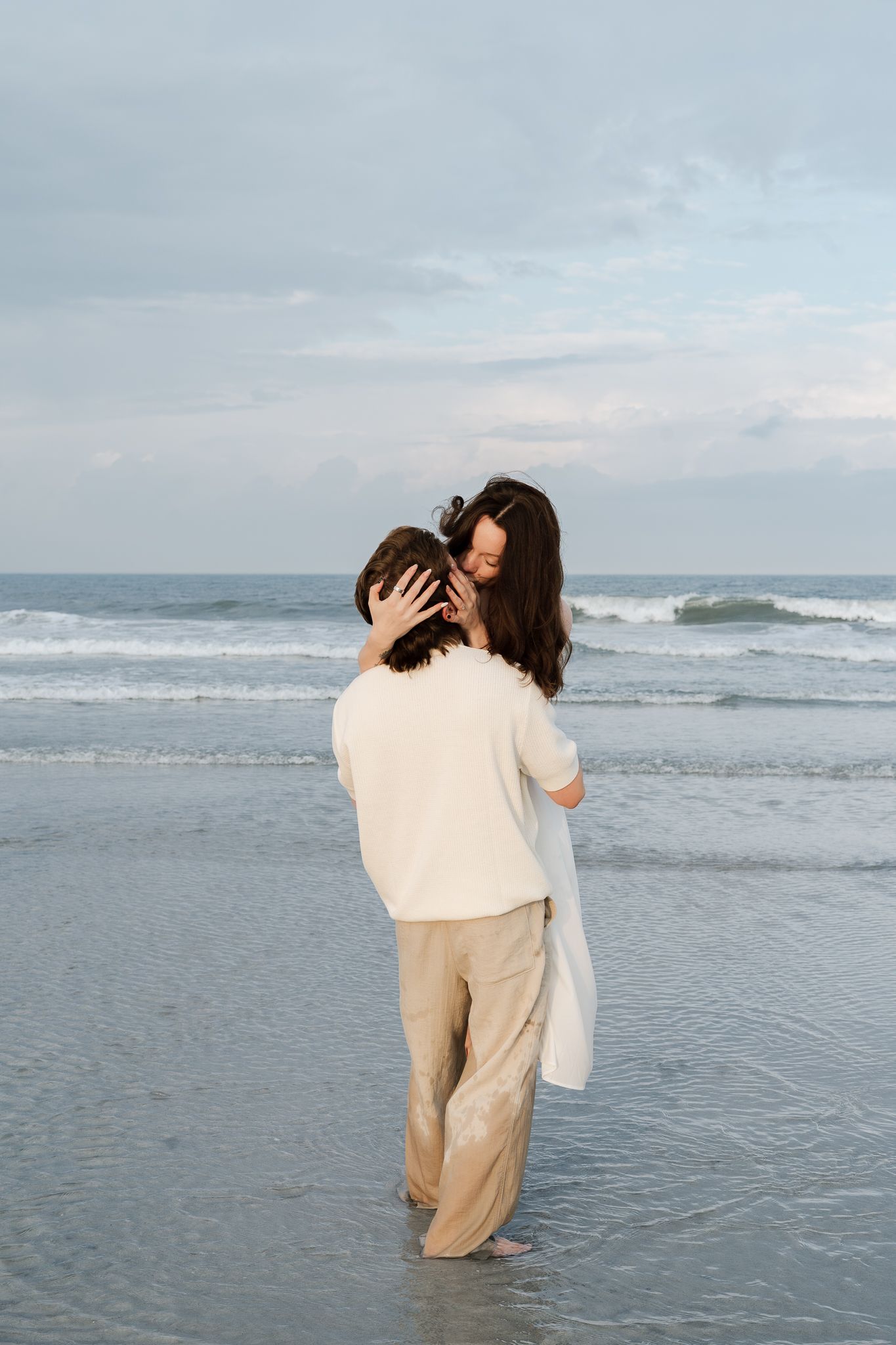 Romantic couple embracing tenderly at the ocean's edge, woman in white top and tan pants with waves behind them.