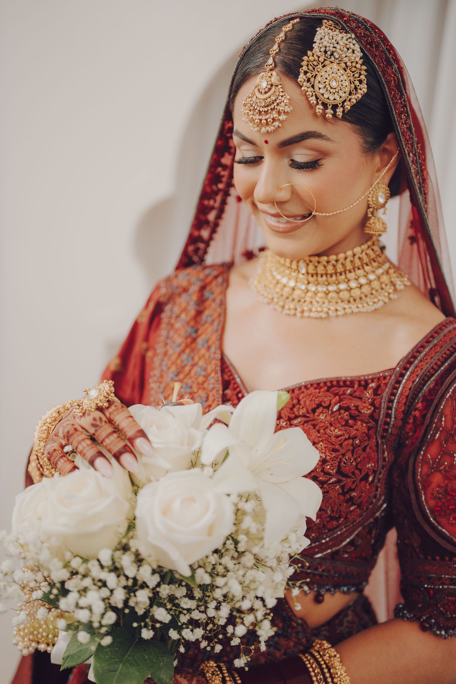 Traditional Indian bridal attire with white rose bouquet and intricate gold jewelry against burgundy lehenga.