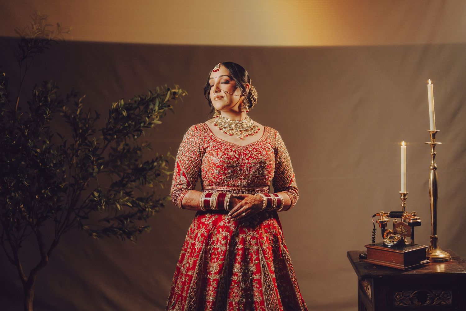 Traditional Indian bride wearing a red embroidered lehenga poses elegantly with candles and decor in moody lighting.