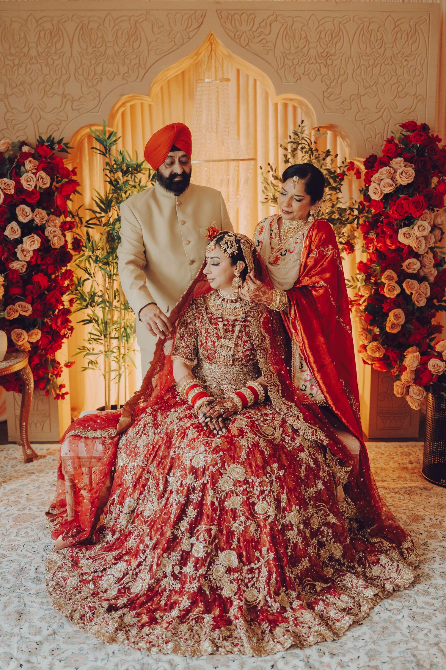 Luxurious Indian wedding scene with ornate red and gold lehenga against floral backdrop with architectural details.