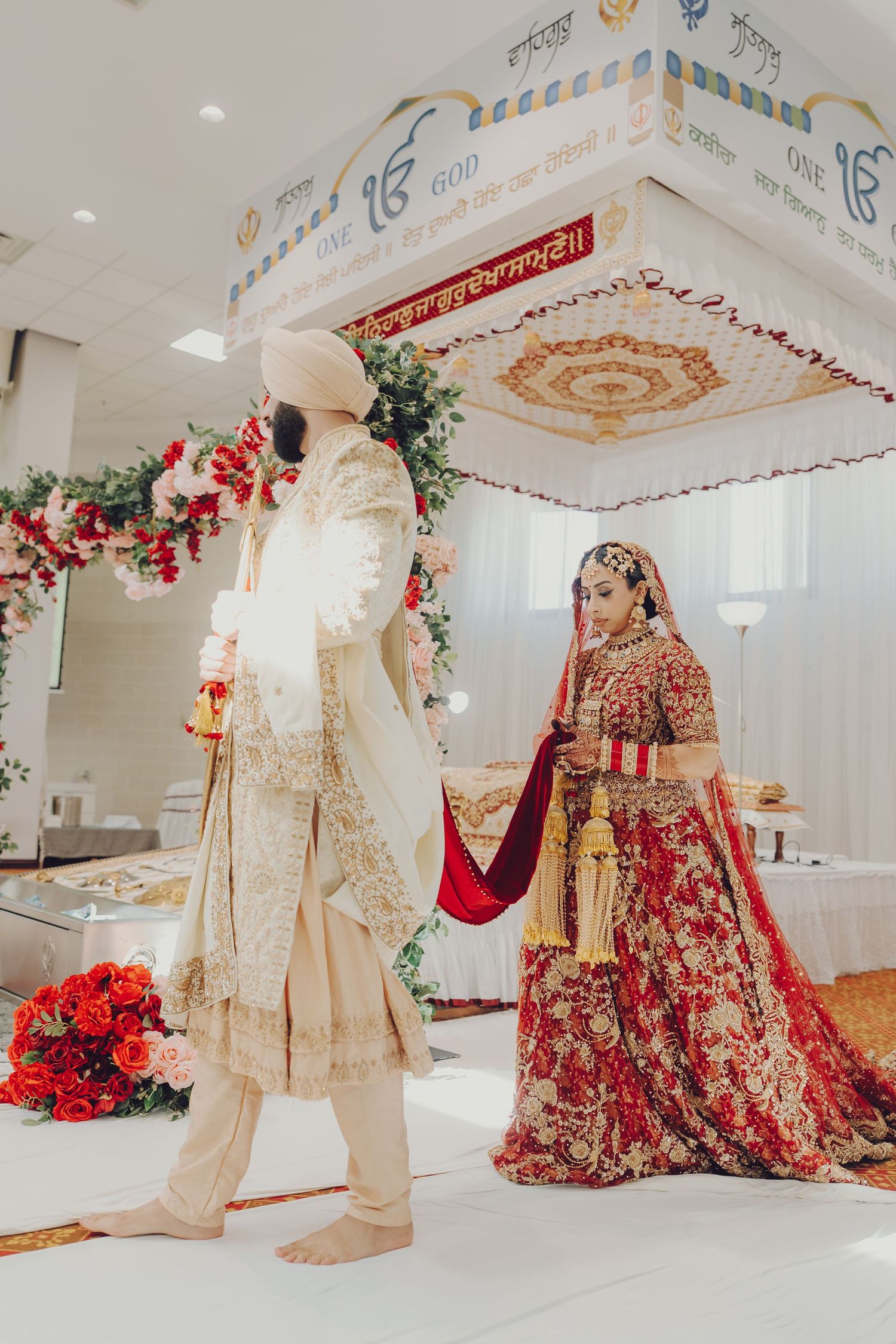 Ornate Sikh wedding venue with decorated ceiling, traditional outfits, and red floral arrangements.