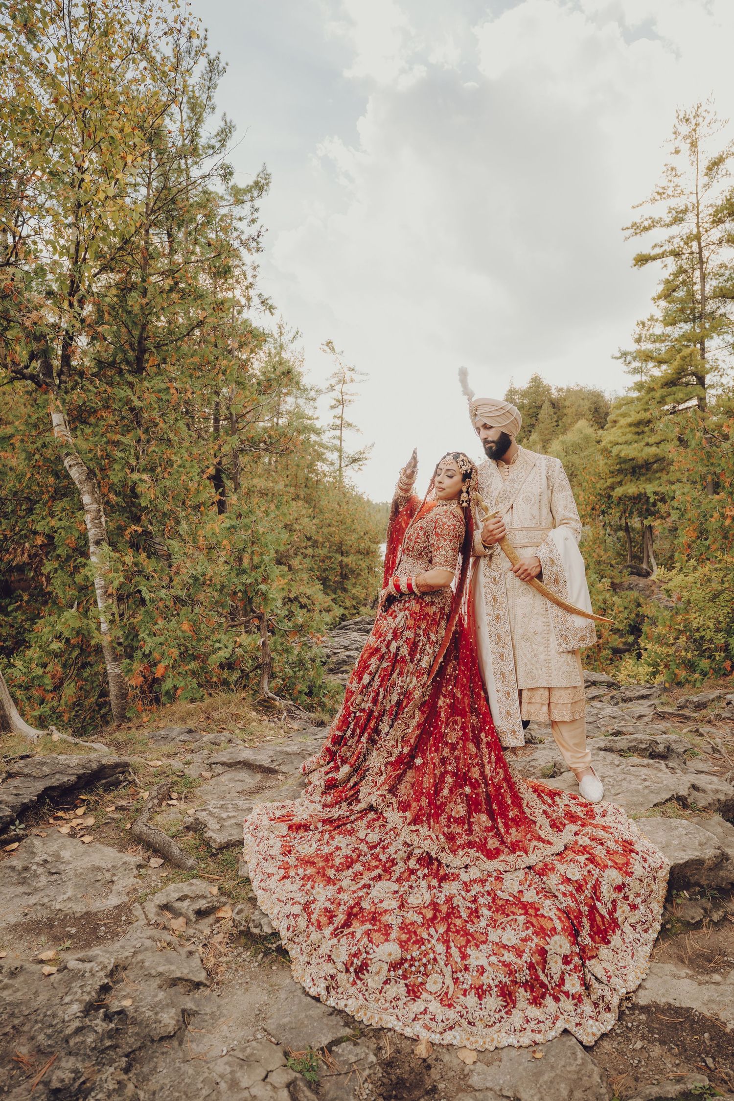 A couple in traditional wedding attire poses on rocky terrain surrounded by autumn trees.