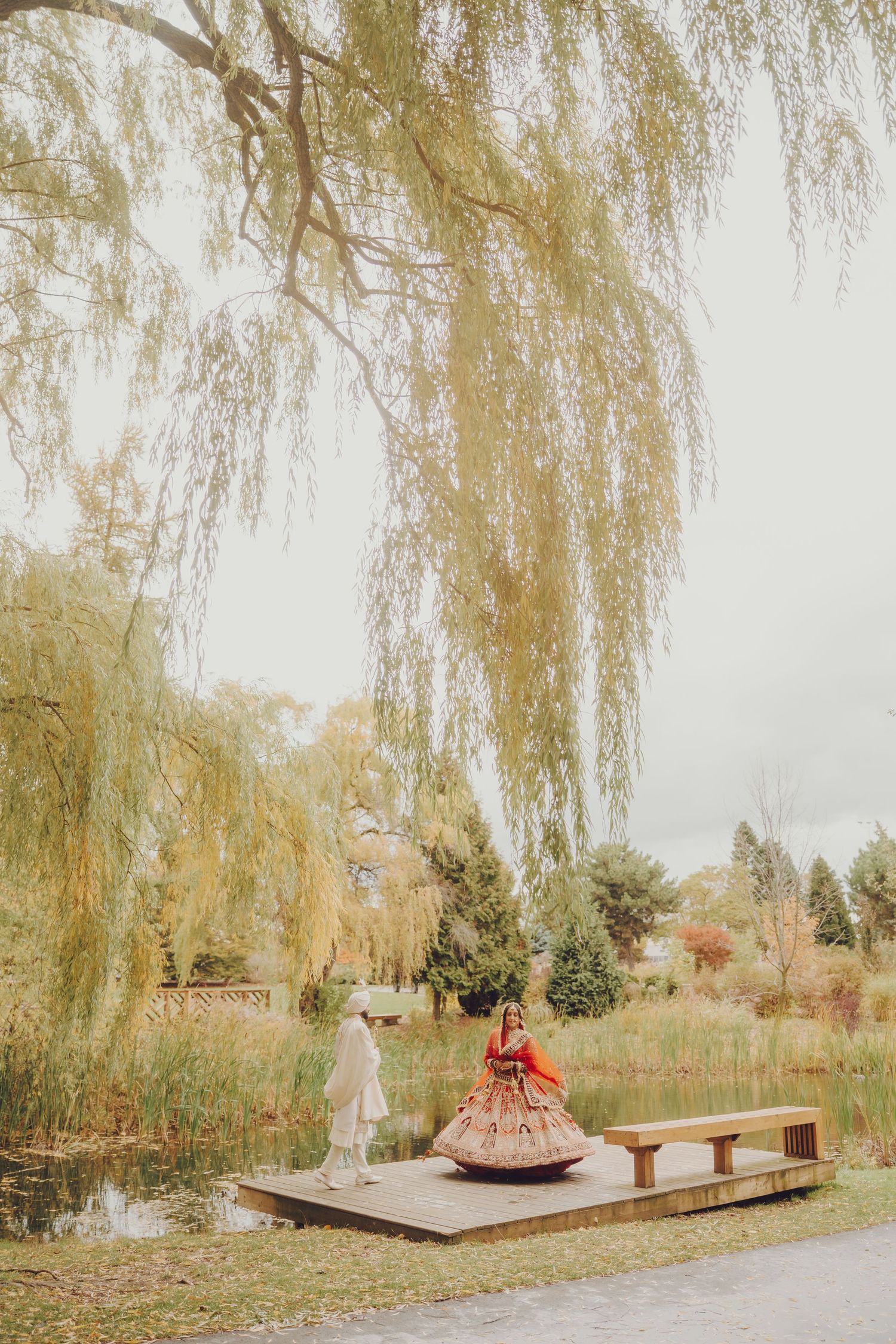 Wedding couple in traditional Indian attire posing on a wooden deck under weeping willow trees.