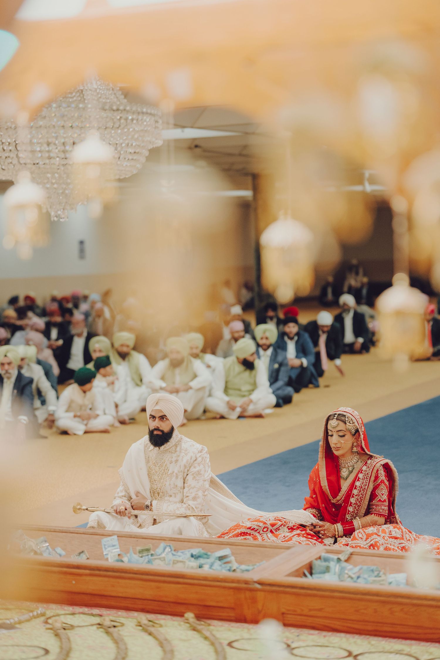 Wedding guests seated on floor during traditional Sikh ceremony with decorative lighting above.