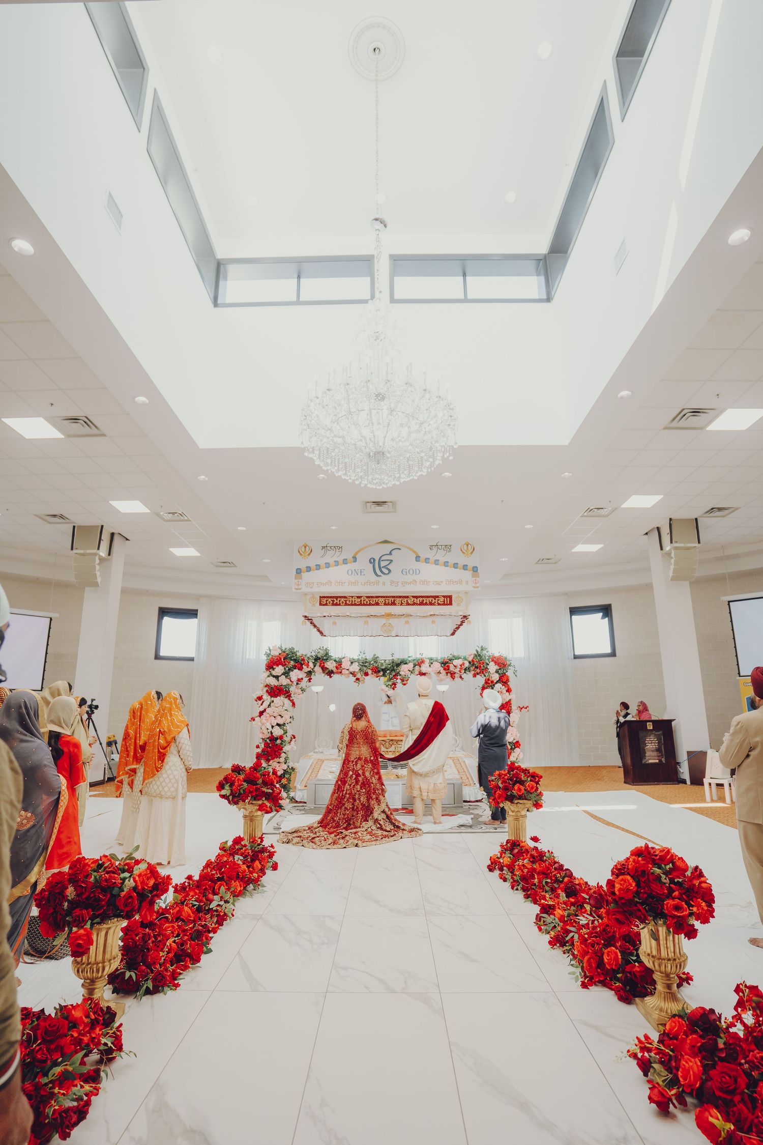 Panoramic view of an elegant wedding venue hall decorated with vibrant red floral arrangements and crystal chandeliers.