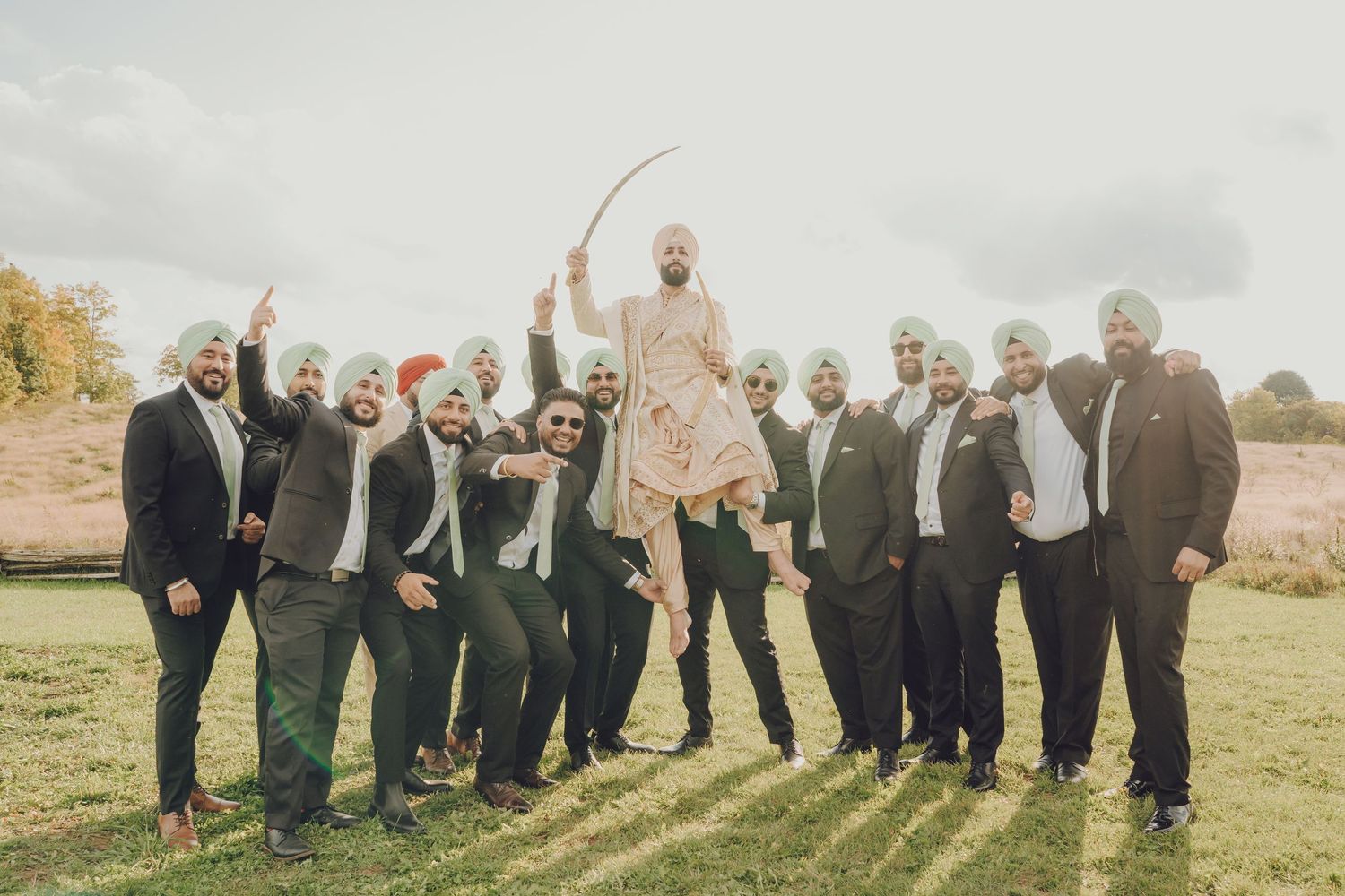 A group of formally dressed people celebrate together outdoors on a grassy field with warm autumn lighting.