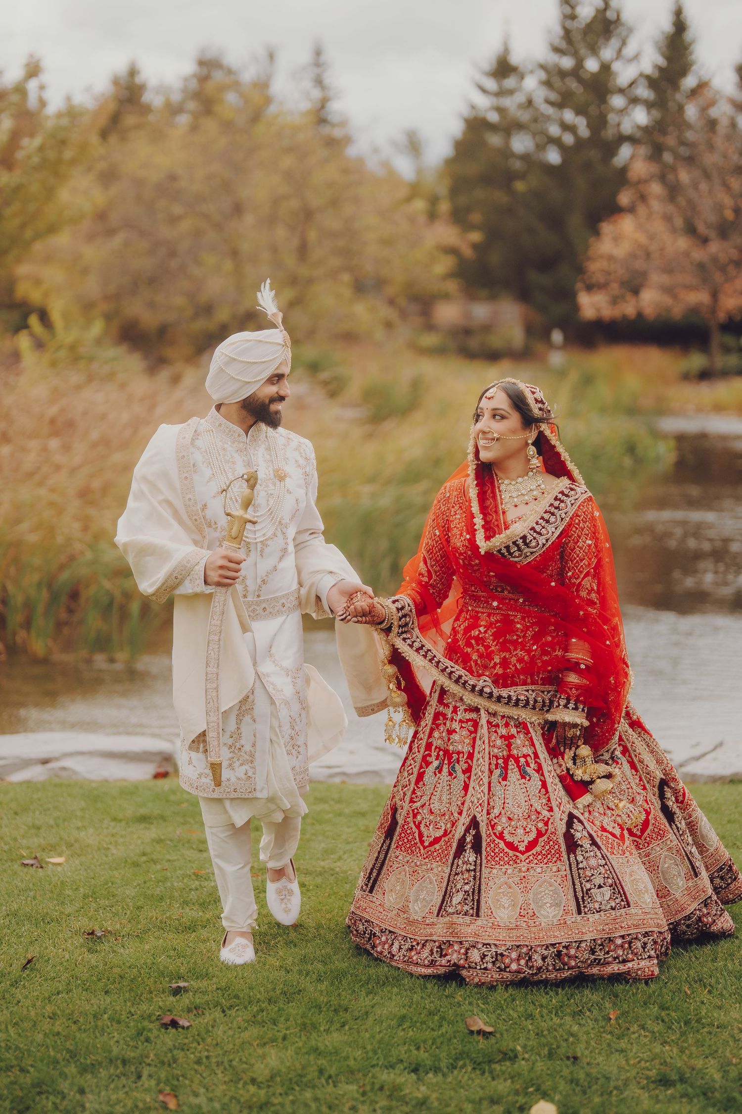 A couple in traditional Indian wedding attire poses outdoors by a lake in autumn with vibrant red lehenga and white sherwani.