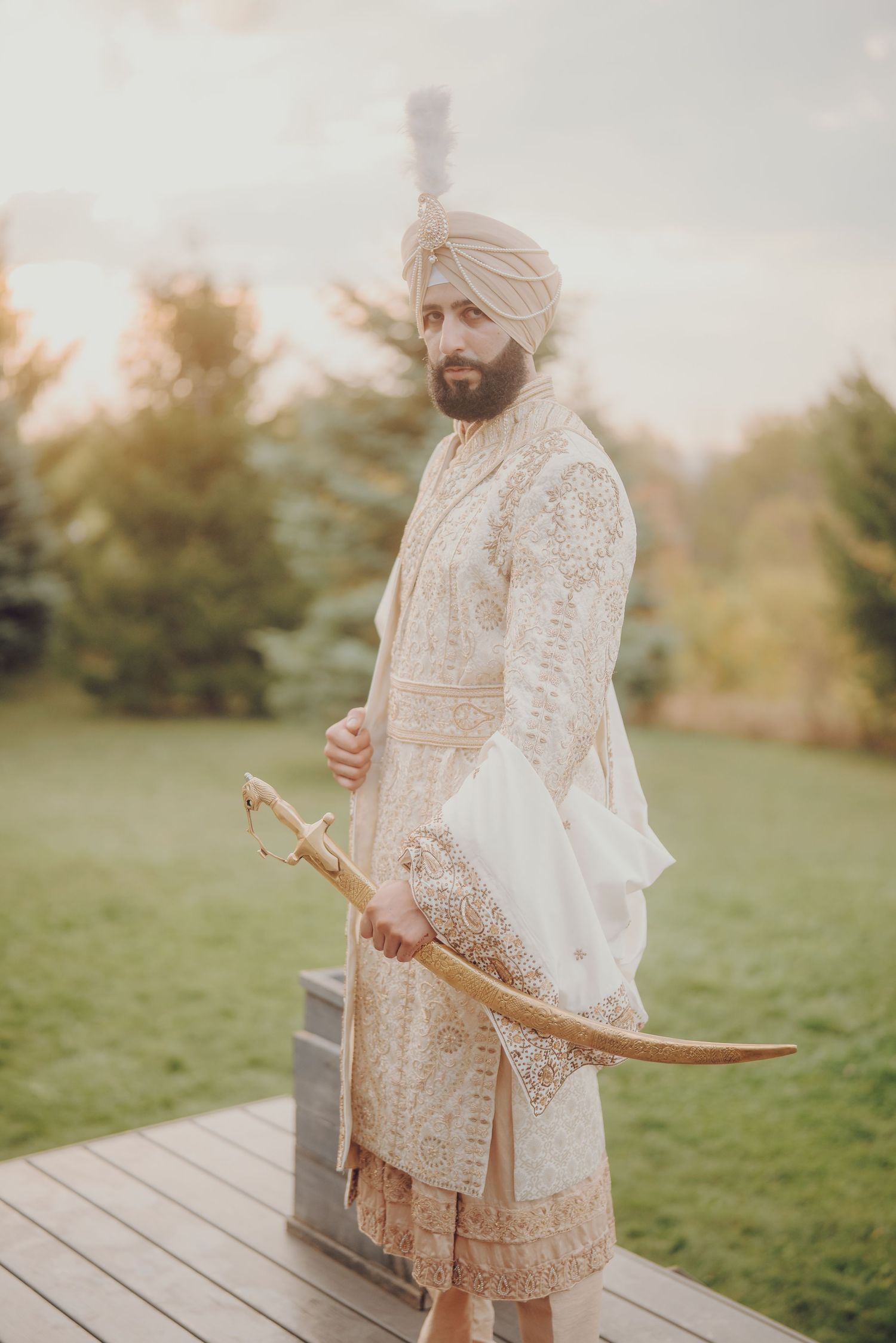 A Sikh groom in cream traditional attire holds a ceremonial sword while standing on a wooden deck outdoors.