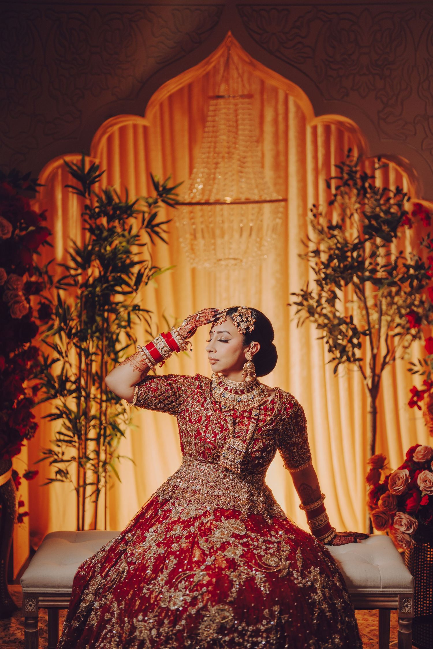 A traditional Indian bride in an ornate red lehenga poses against a golden archway backdrop with decorative plants.