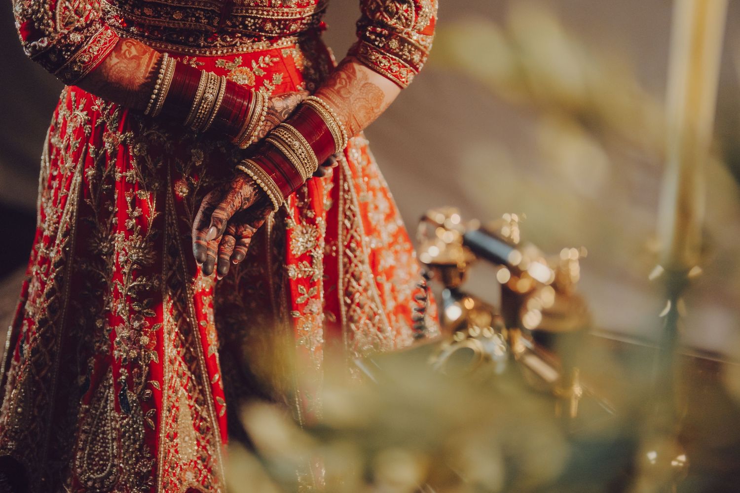 Close-up of hands adorned with traditional Indian bridal mehndi and bangles against a red embroidered dress.