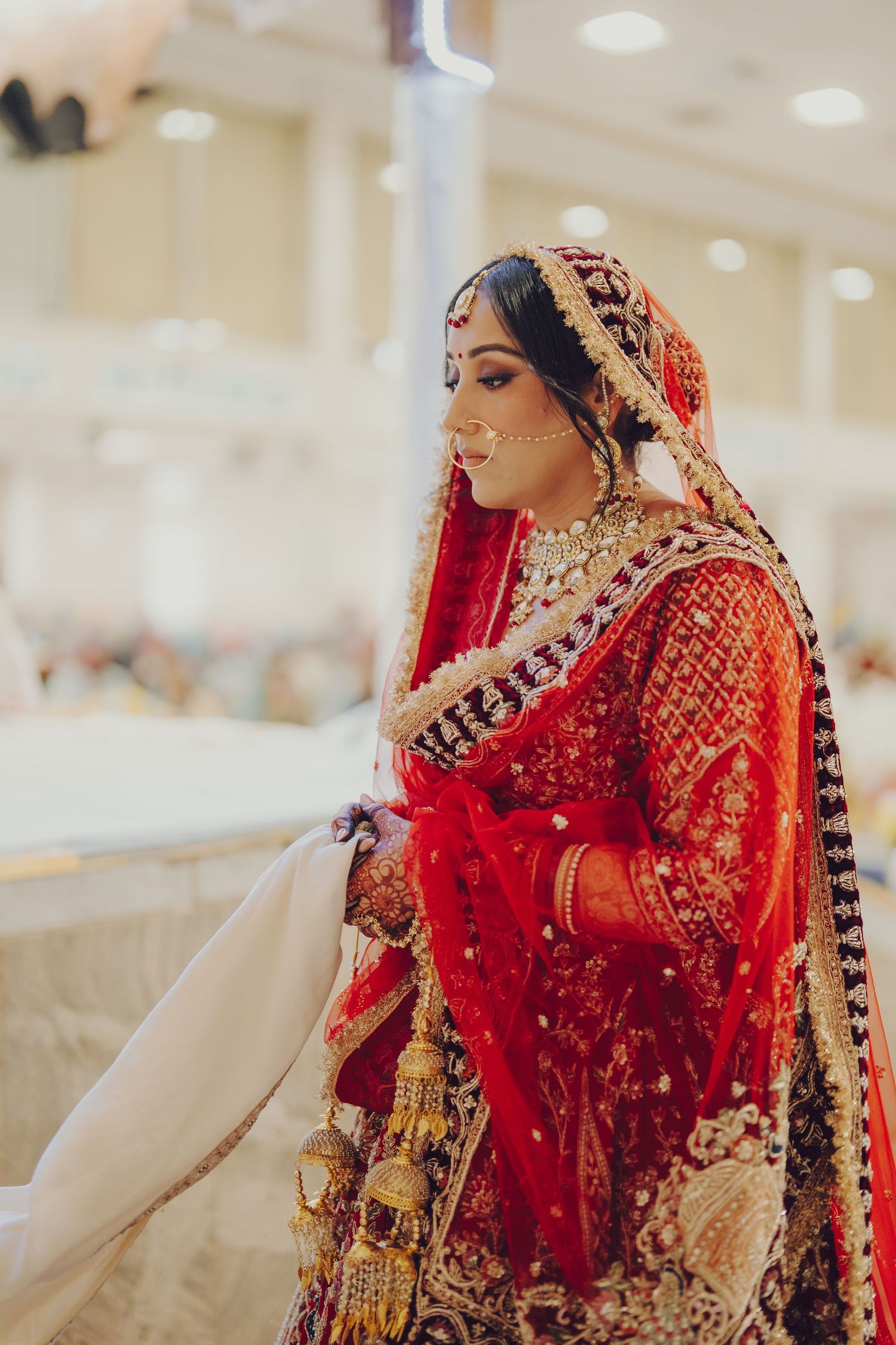 A bride in a stunning red and gold embroidered lehenga with traditional Indian bridal jewelry poses elegantly.