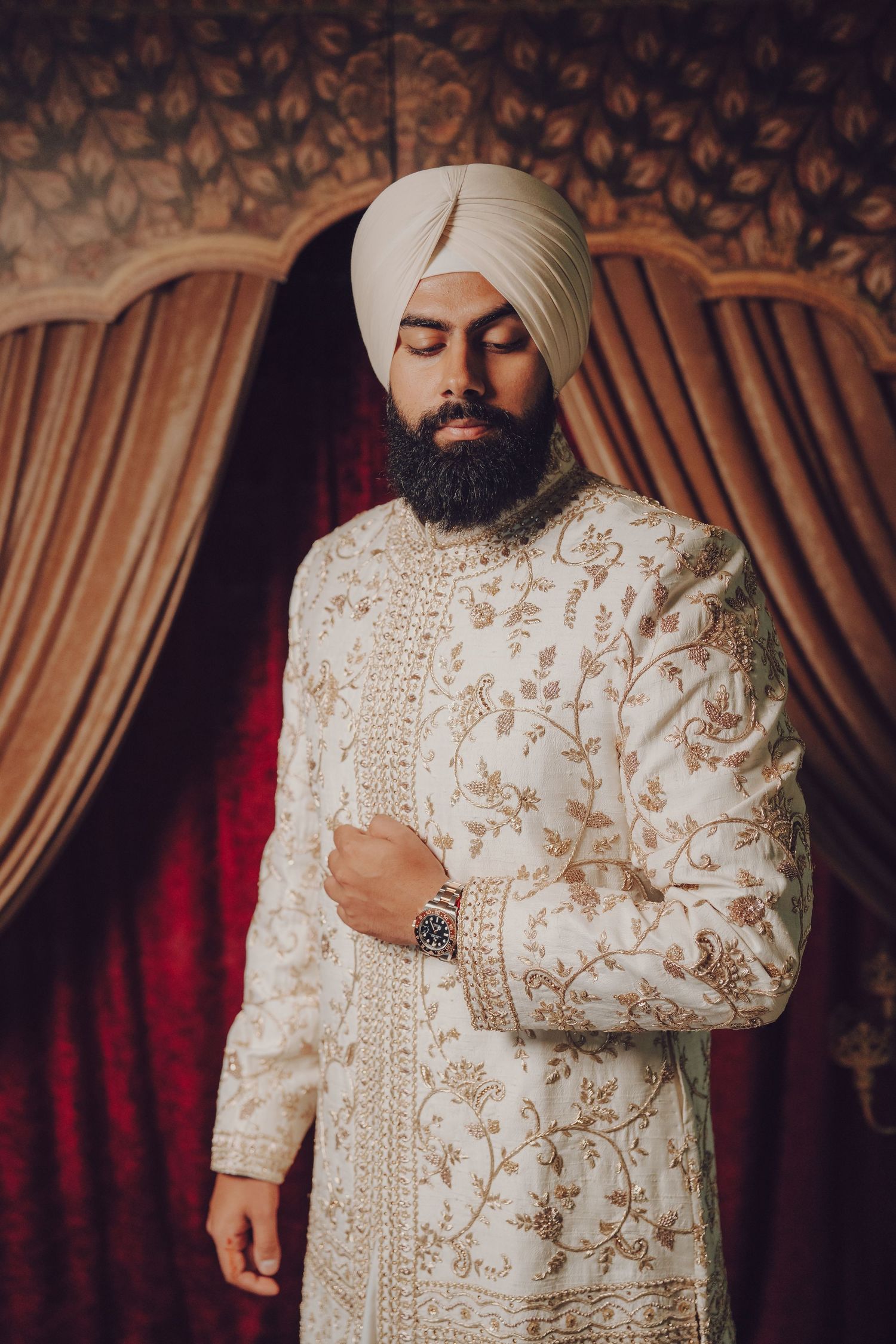 A groom in a cream-colored sherwani and turban poses against an ornate backdrop.