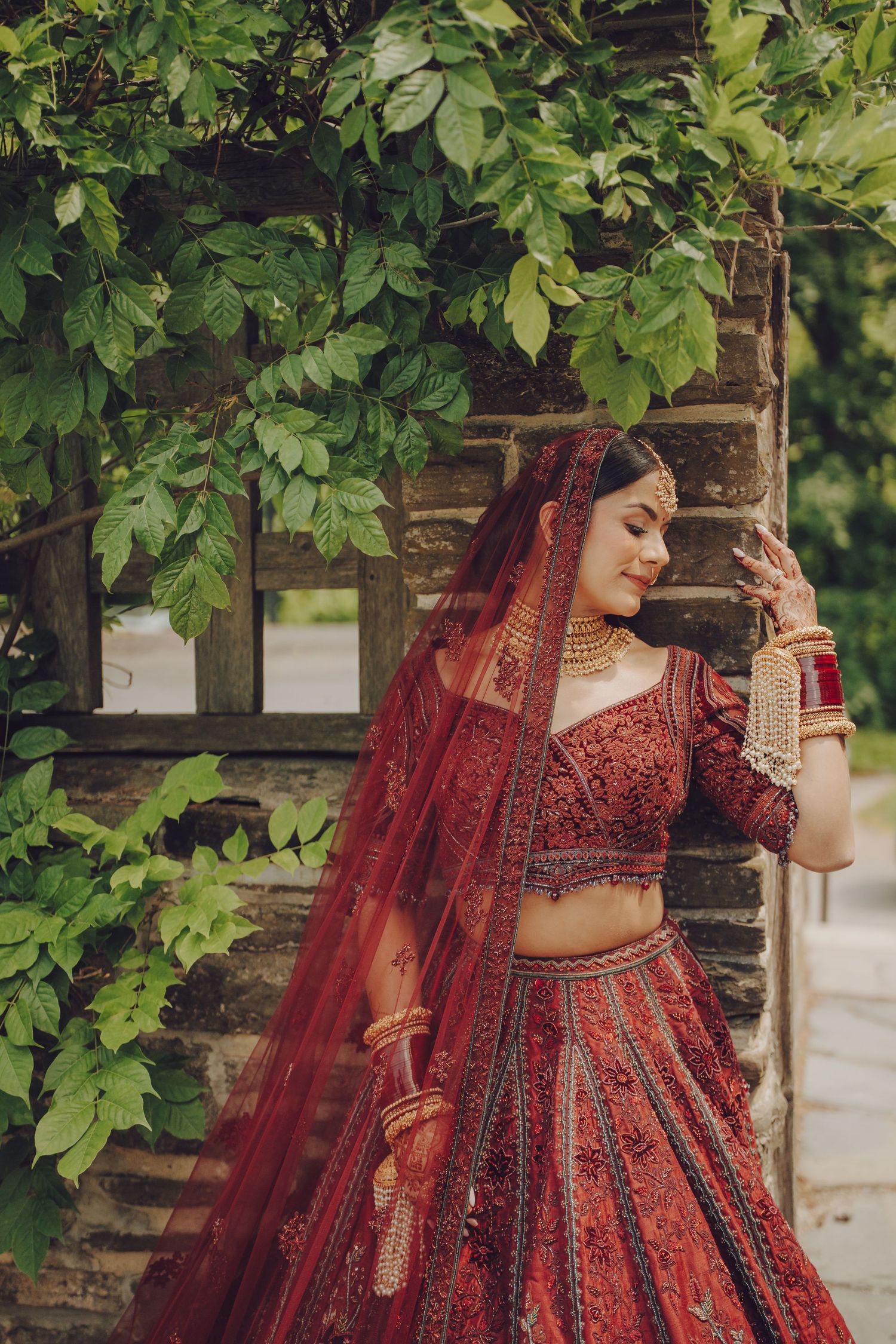 Artistic bridal poses against lush green foliage wearing a deep red embellished lehenga.