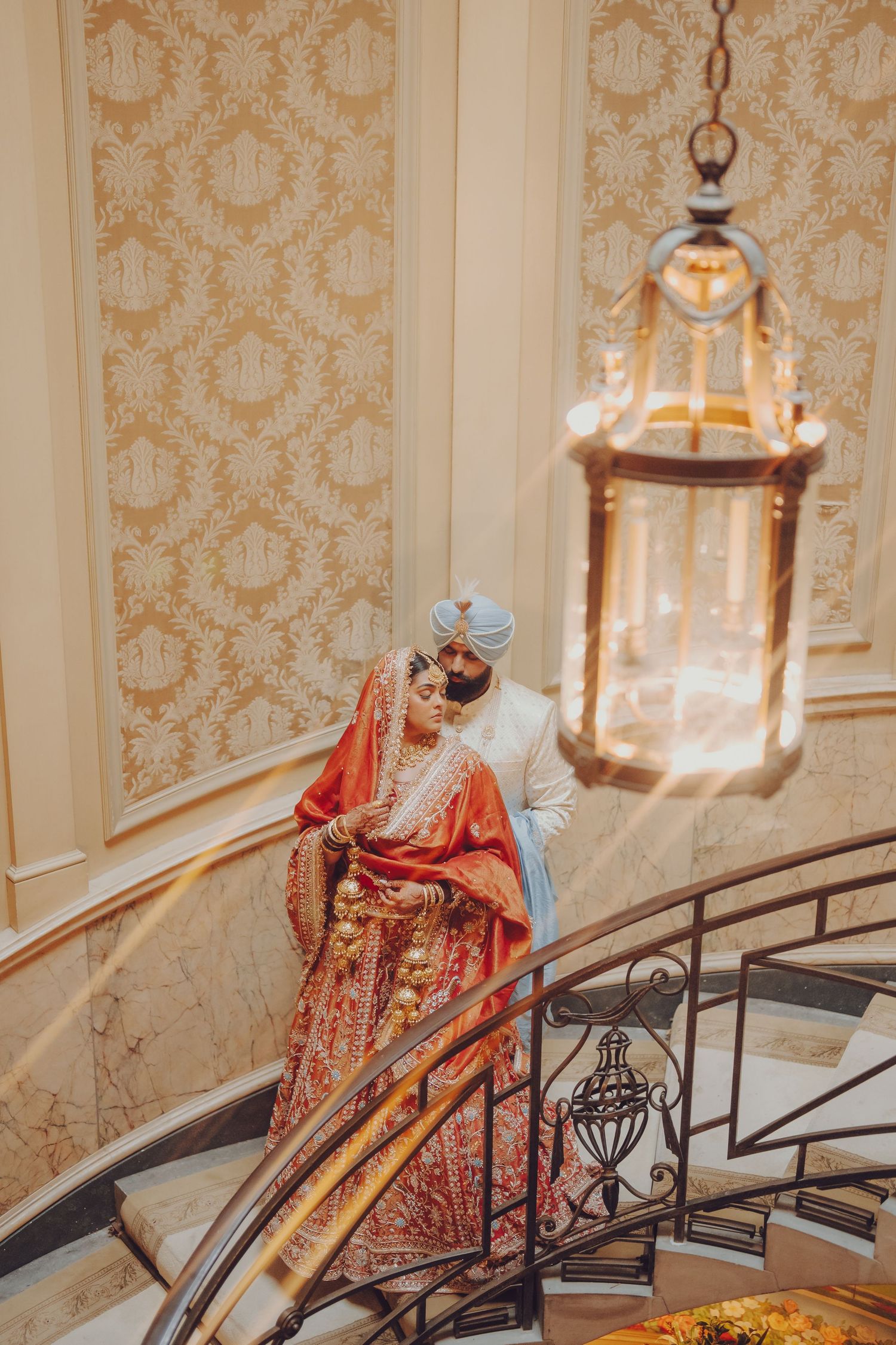 Elegant couple in South Asian wedding attire poses on grand staircase with ornate wallpaper.