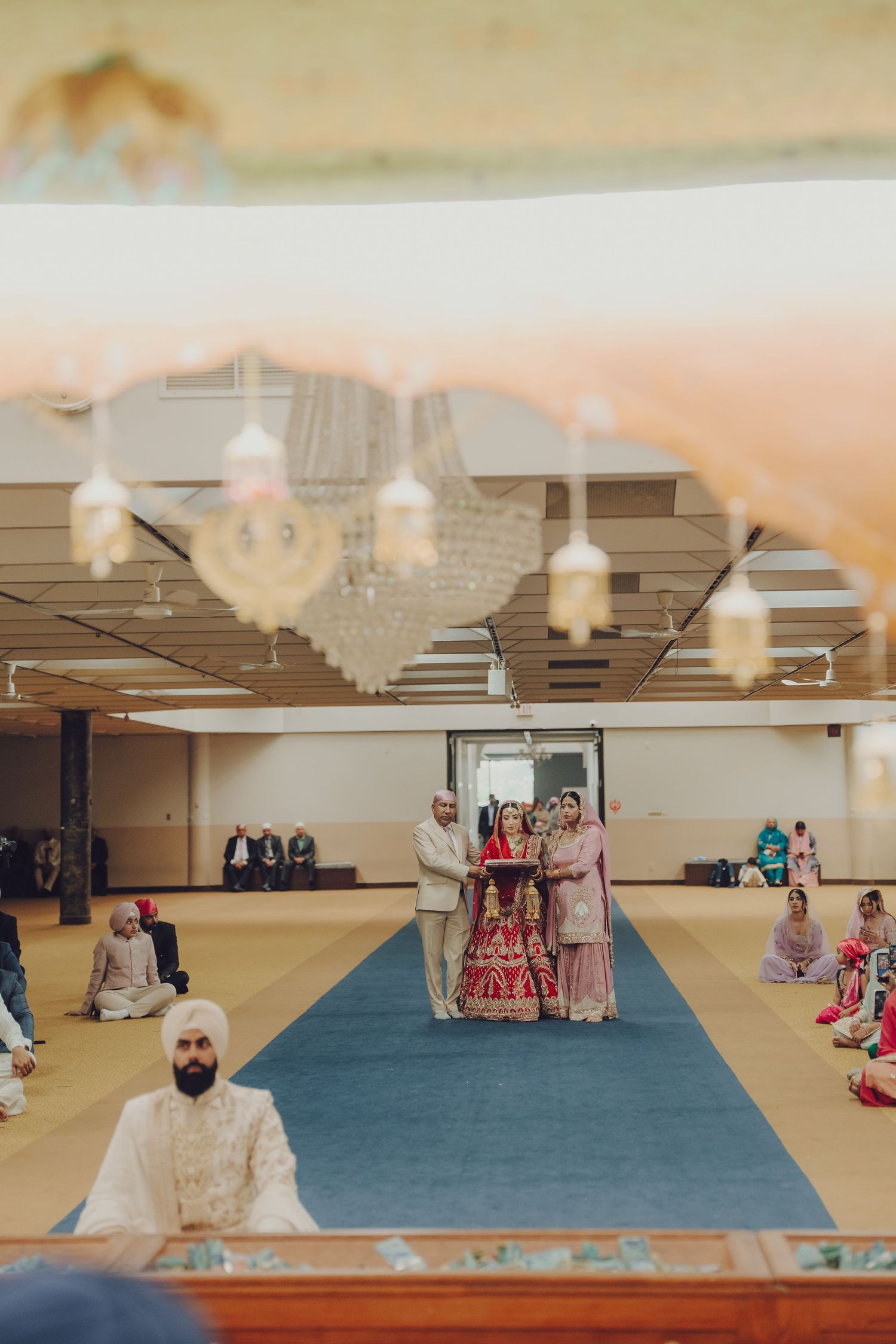 A wide-angle shot of a Sikh temple interior with blue carpeting and hanging pendant lights during a ceremony.