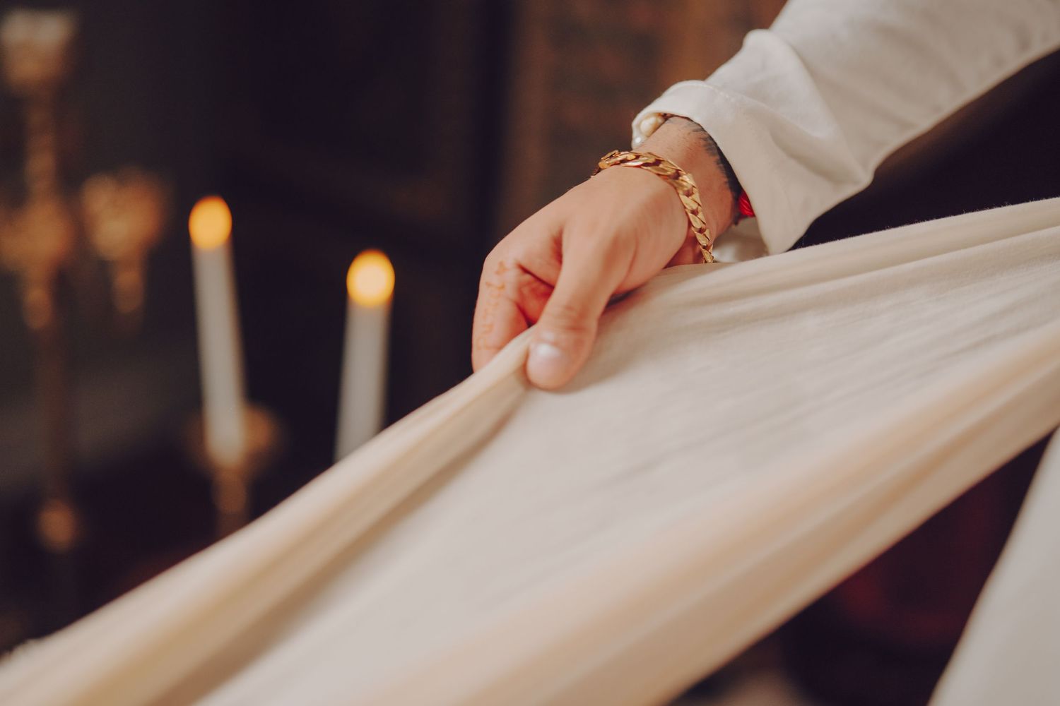 Hands carefully folding white fabric on a dark wooden surface with warm candlelight in the background.