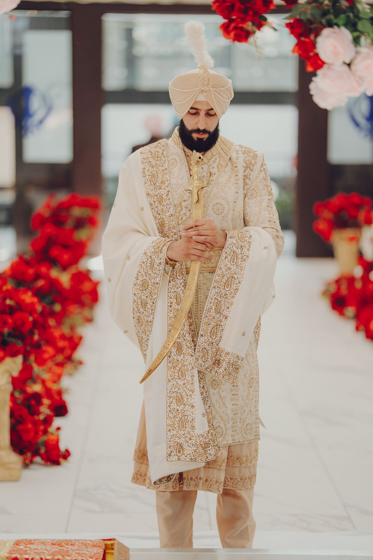 Groom in elegant cream and gold embroidered sherwani poses against backdrop of vibrant red flowers at Indian wedding.