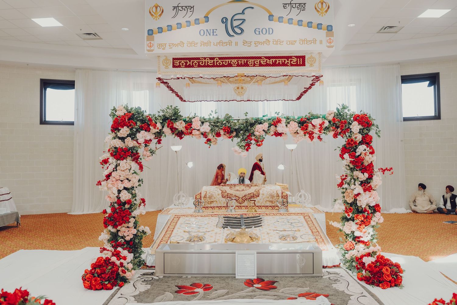 Sikh wedding ceremony setup with white mandap decorated with red roses and floral arch in a temple hall.