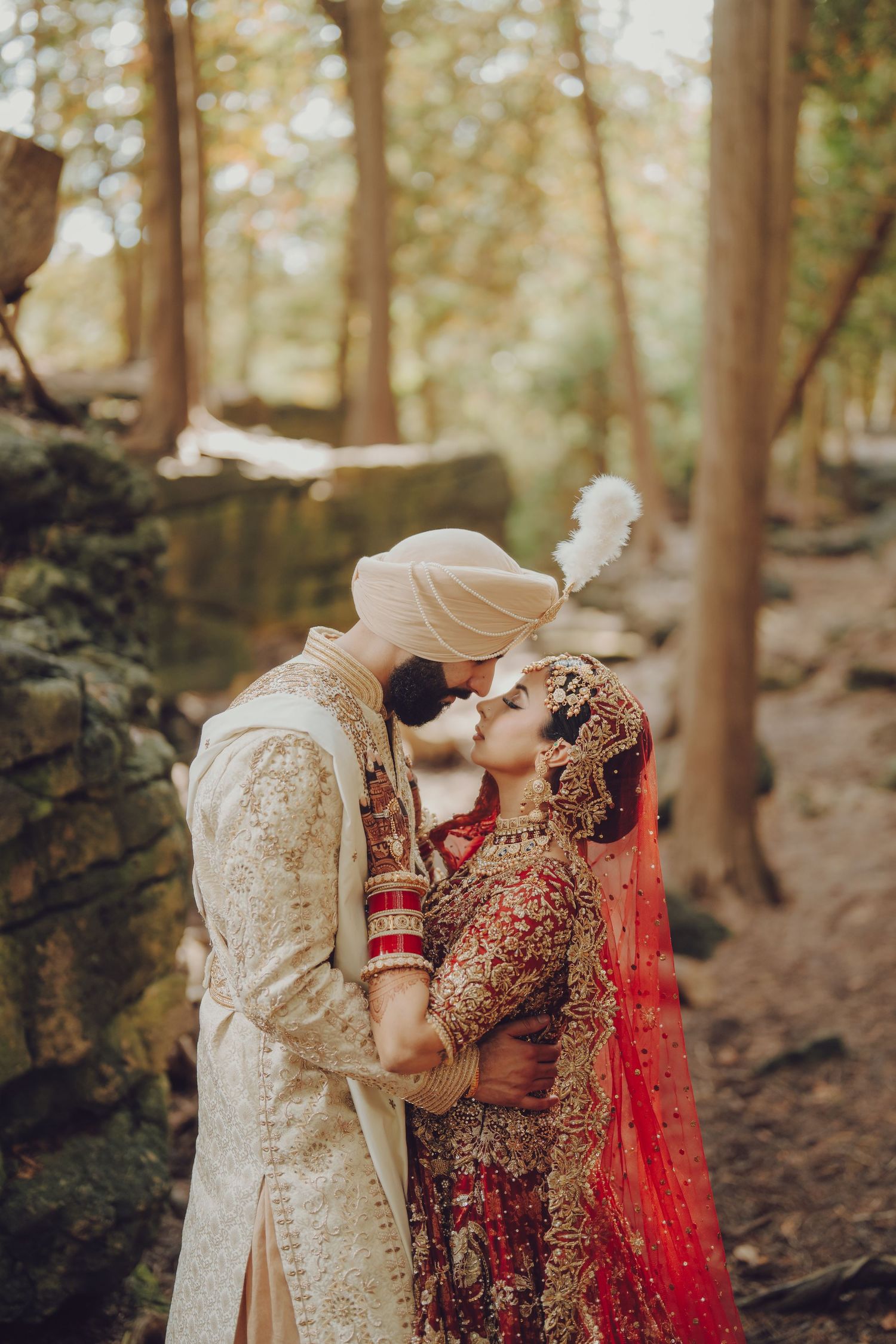 Traditional Indian wedding couple embraces in a romantic forest setting wearing ornate cream and red wedding attire.