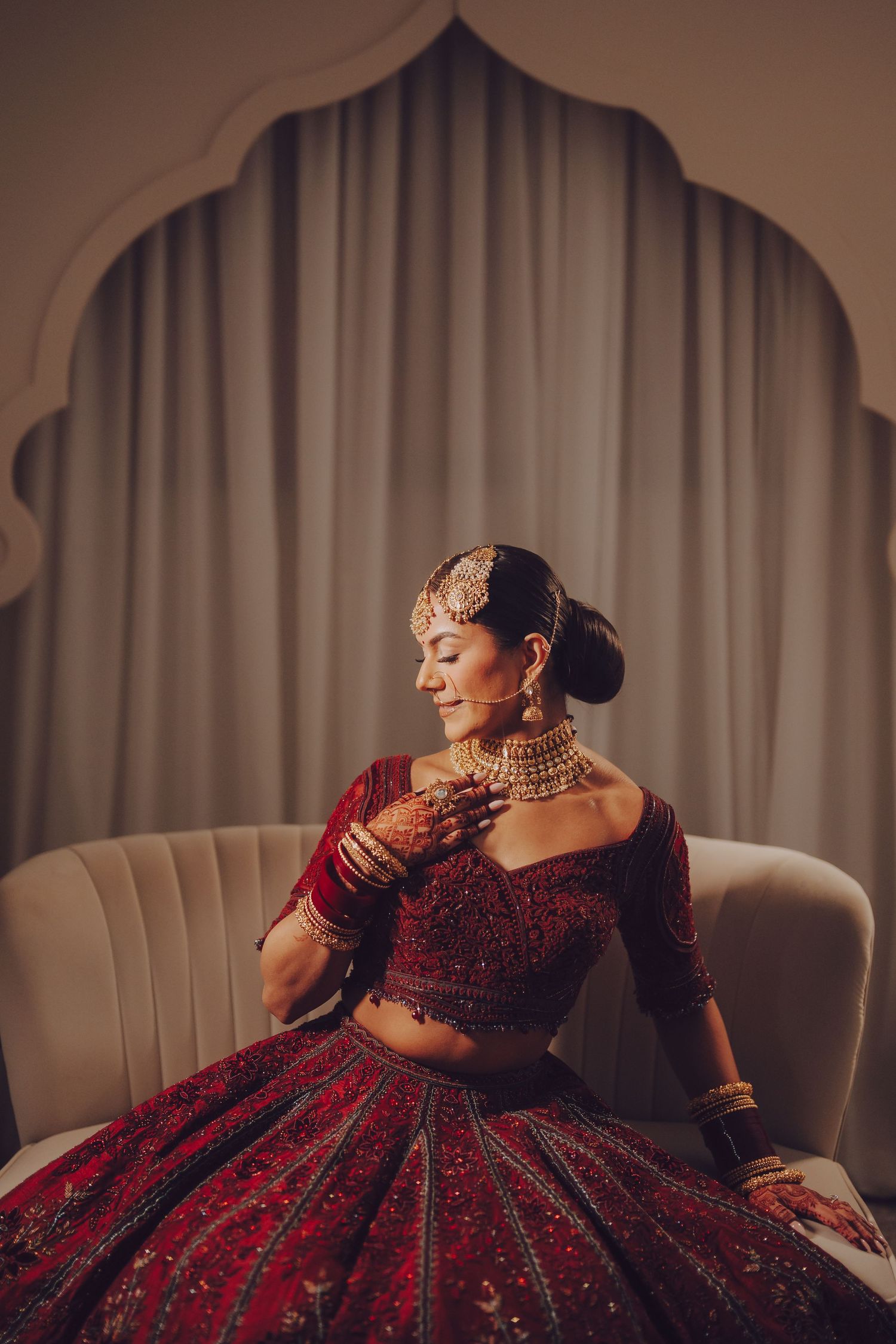 A bride in a deep red velvet lehenga poses elegantly on a cream colored chair during a wedding photoshoot.