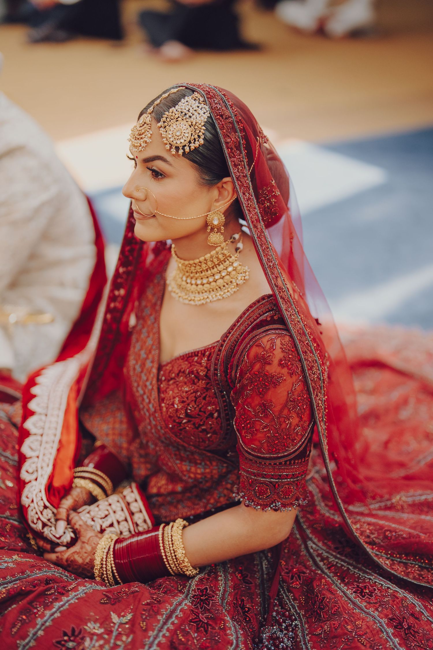 Bride in deep red embellished lehenga with traditional Indian bridal jewelry and makeup.