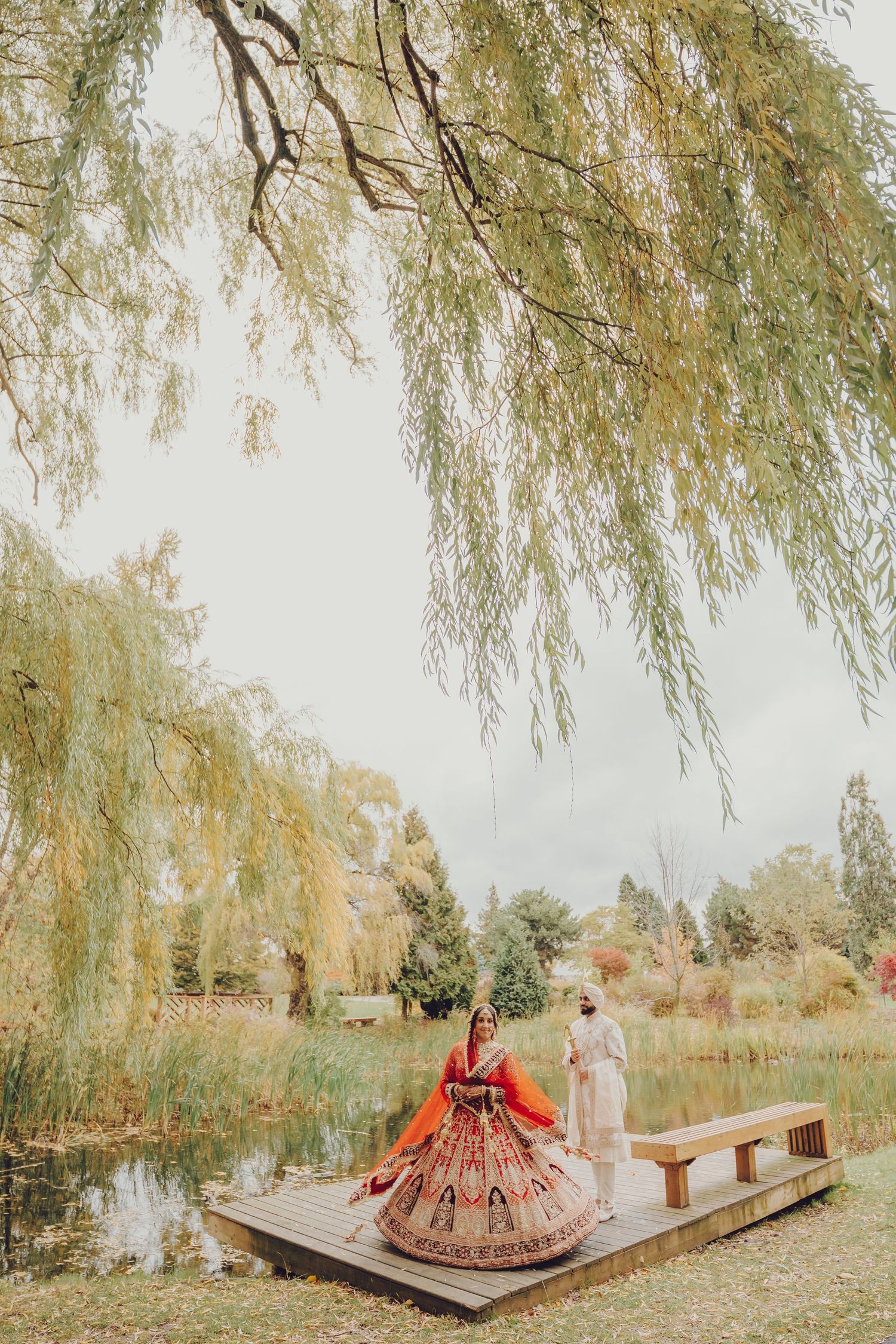 Wedding couple on wooden dock under weeping willow tree by serene lake.