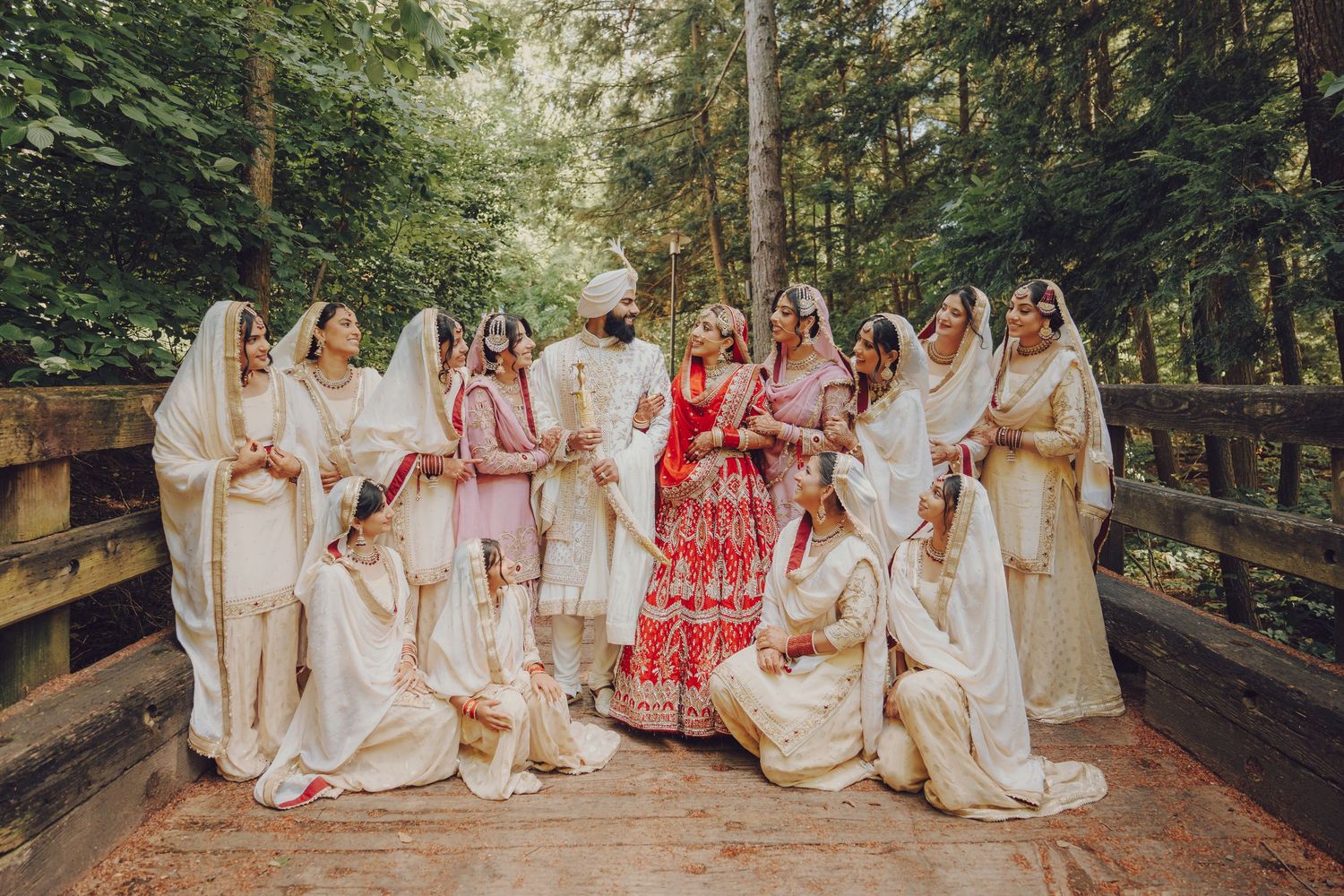 A wedding party in coordinated white and red outfits poses on a wooden bridge in a forest setting.