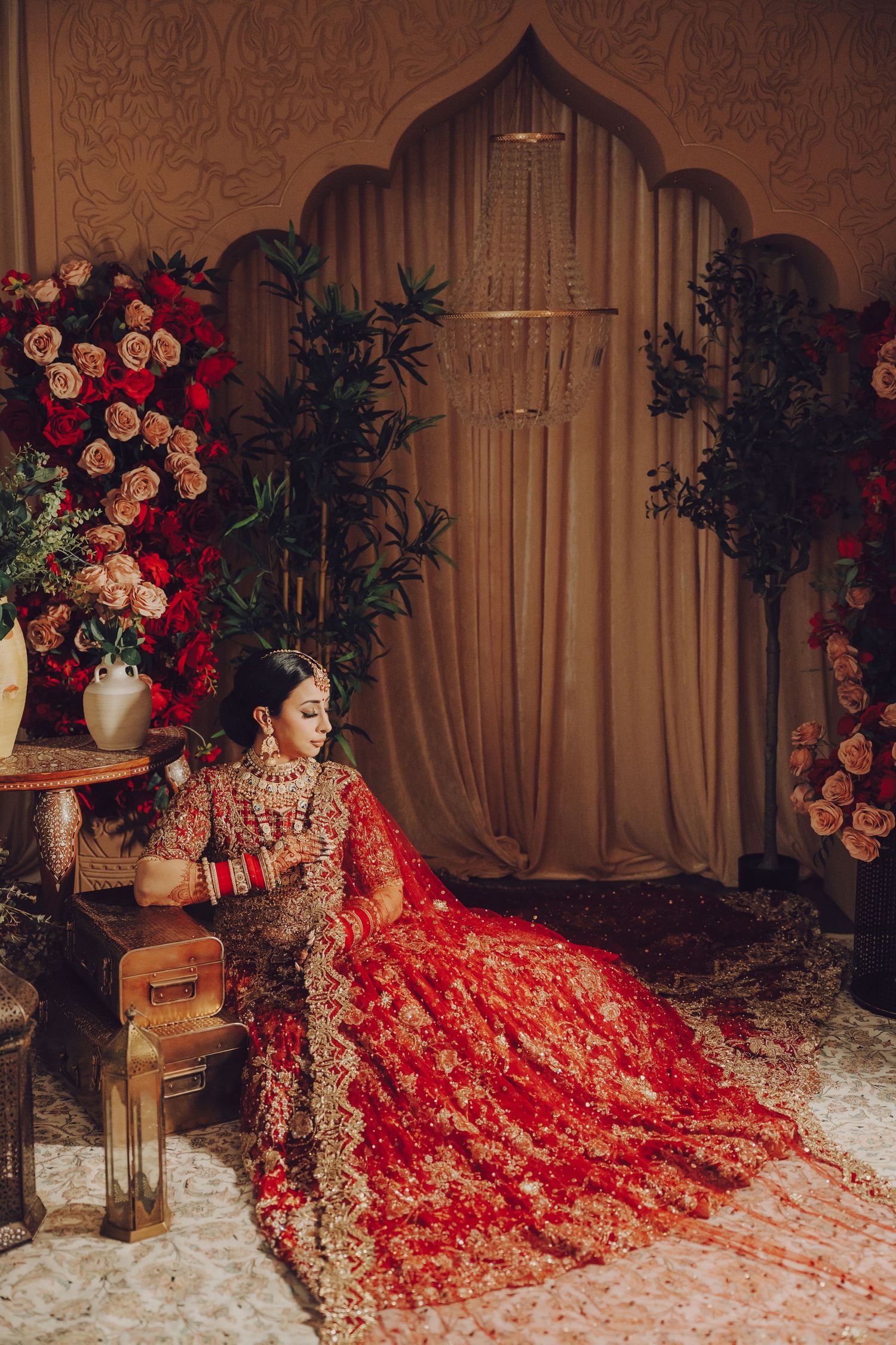 Traditional South Asian bride in ornate red lehenga poses elegantly against floral backdrop with arch and drapes.