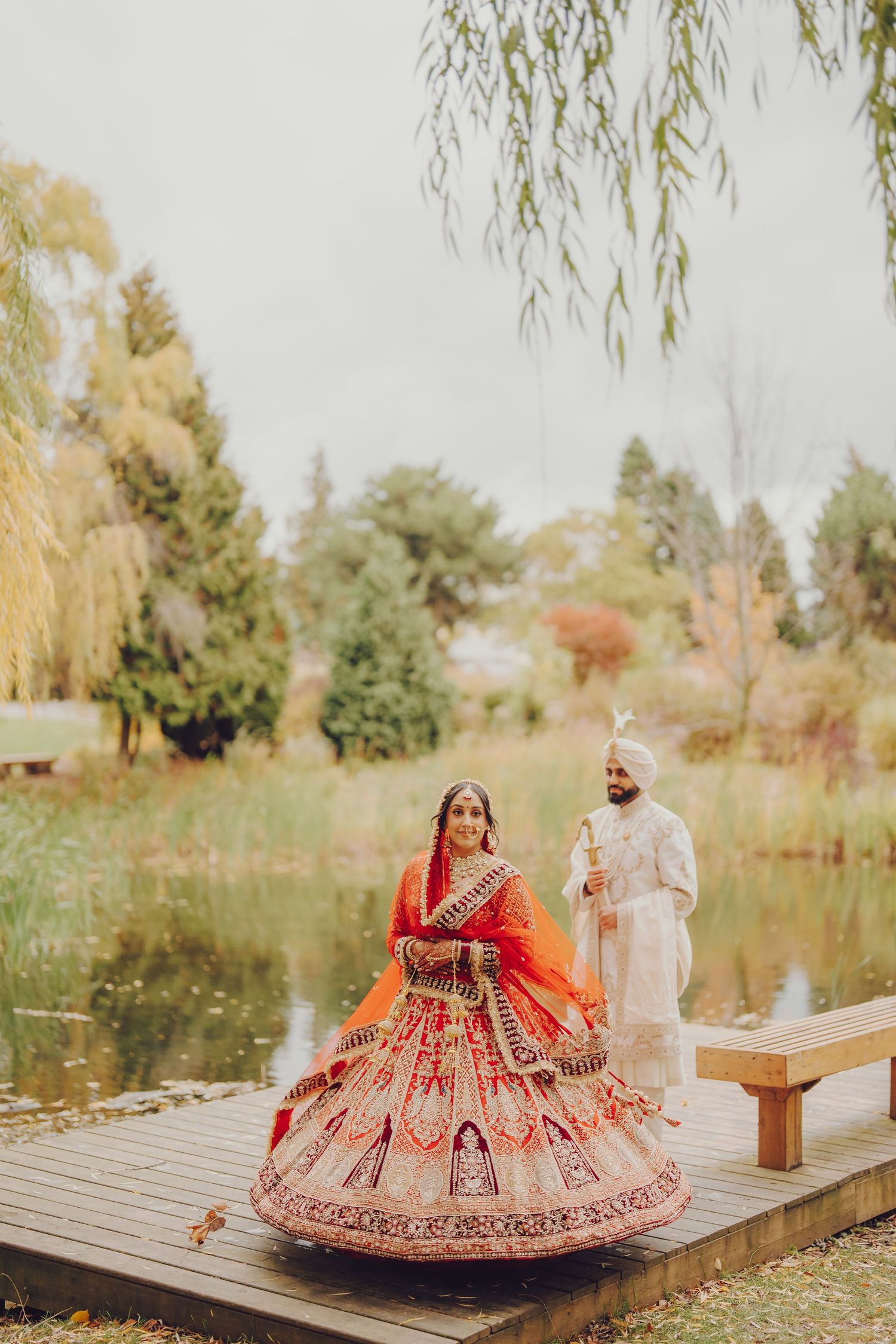 Couple in traditional Indian wedding attire posing on wooden dock by autumn pond.